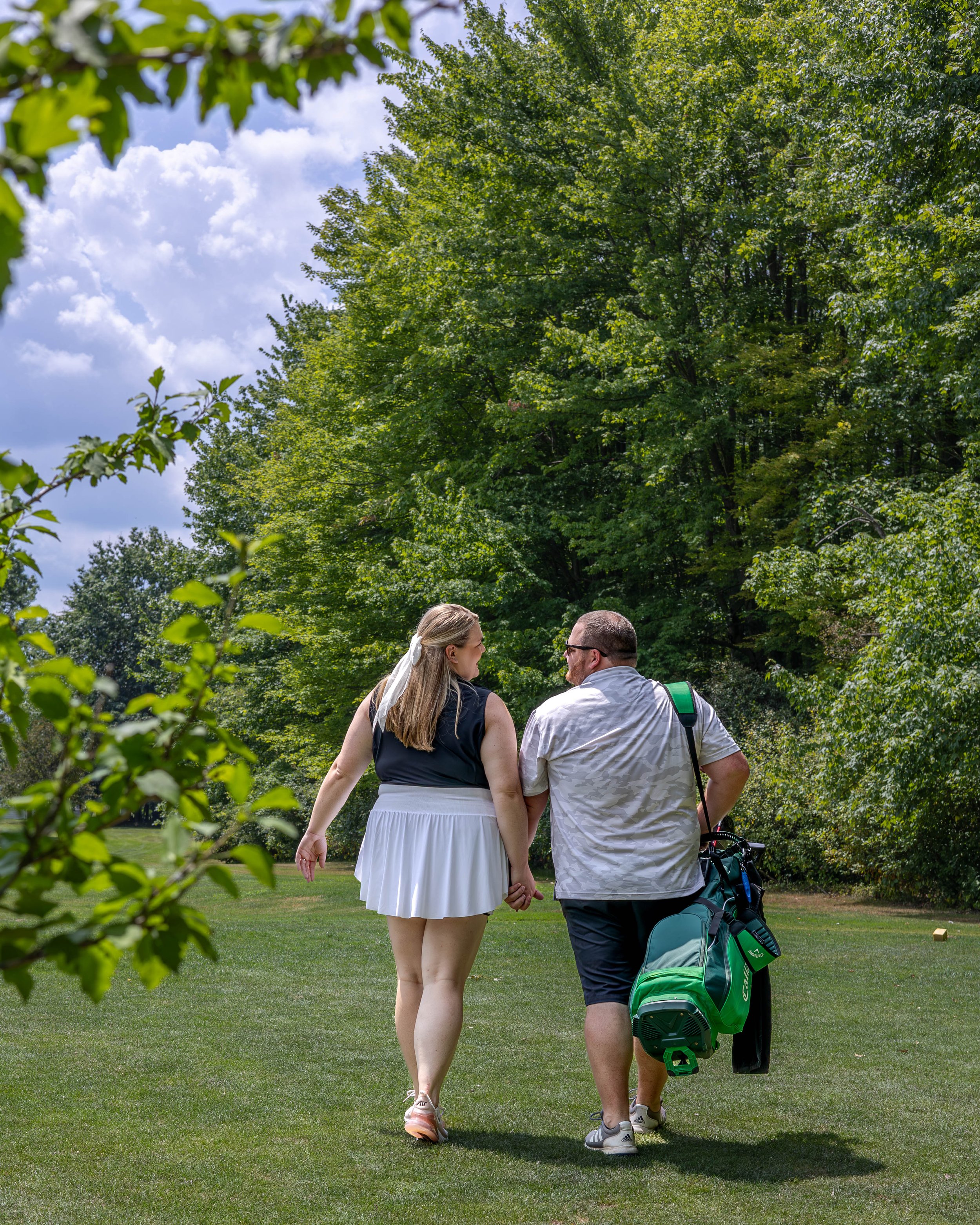 A couple holding hands while walking on a golf course with greenery and trees in the background