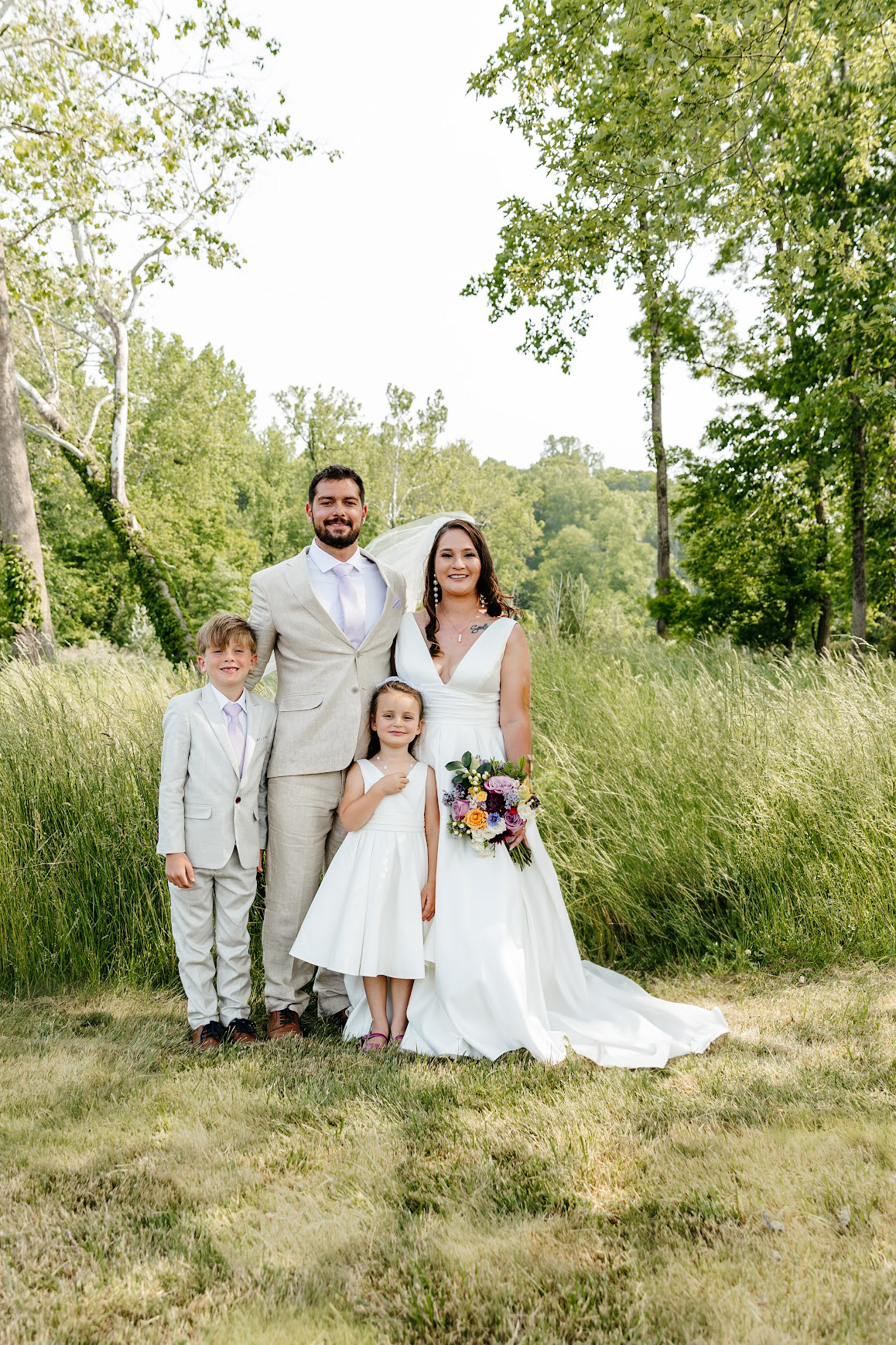 A wedding couple with two children standing outdoors on grass in a green, wooded area. The groom is wearing a beige suit, and the bride is in a white wedding dress holding a bouquet. A young boy and girl are in white outfits. The group smiles at the camera with trees and greenery in the background.