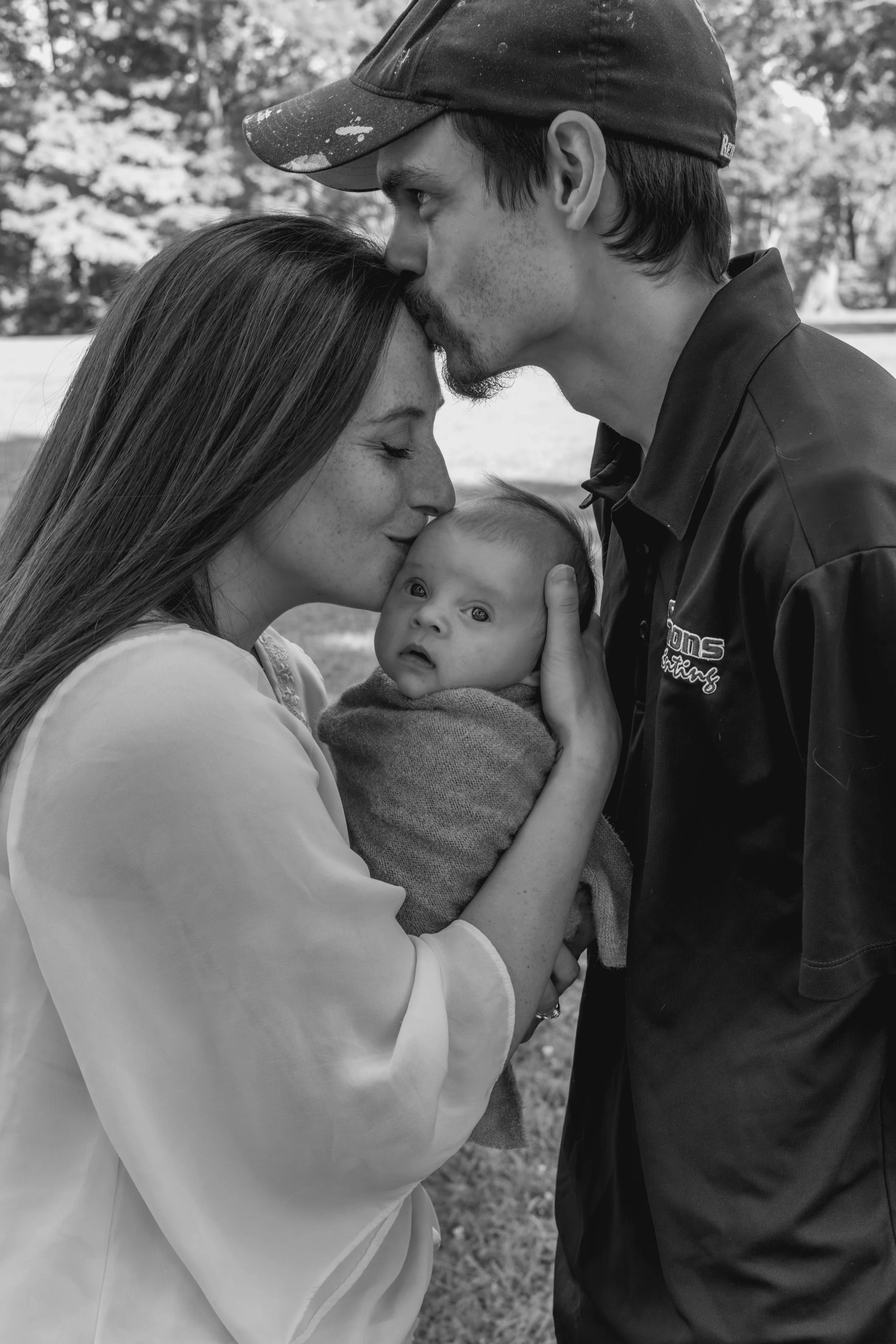 A black and white photo of a woman with long hair and a man with a baseball cap kissing a baby on the forehead outdoors, with trees in the background.
