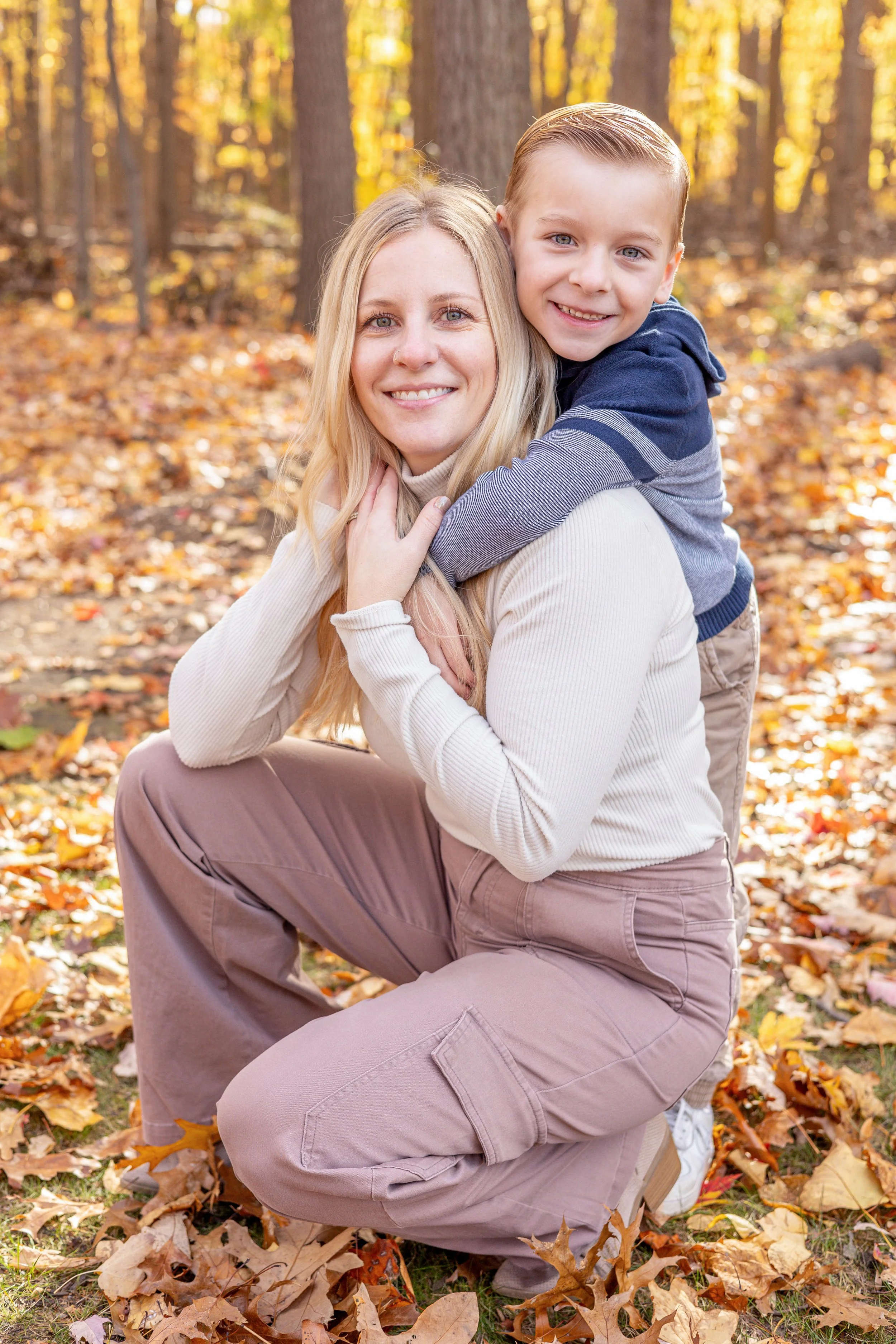 A woman and a young boy smiling together outdoors during fall, surrounded by fallen leaves and trees with autumn foliage. The boy is on the woman's back, hugging her, both dressed casually.