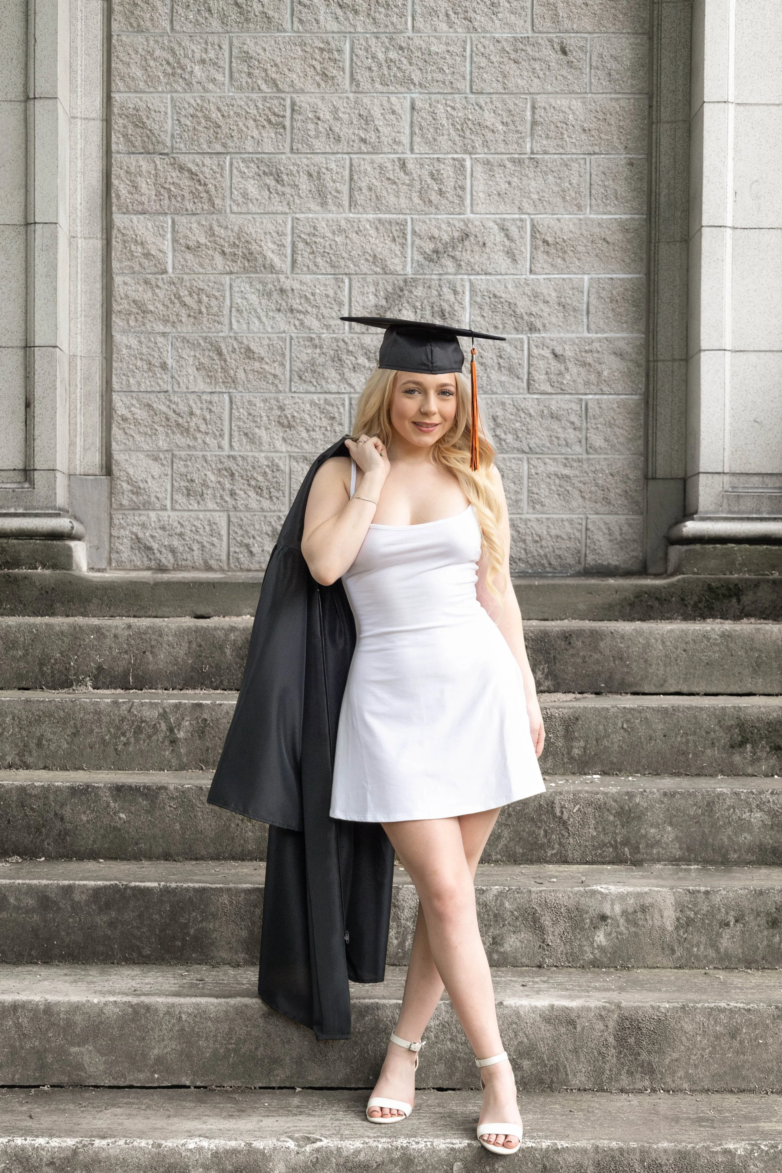 A young woman with long blonde hair wearing a white dress, white high heels, and a black graduation cap with a tassel, standing on concrete steps in front of a stone wall, holding a black graduation gown over her shoulder.