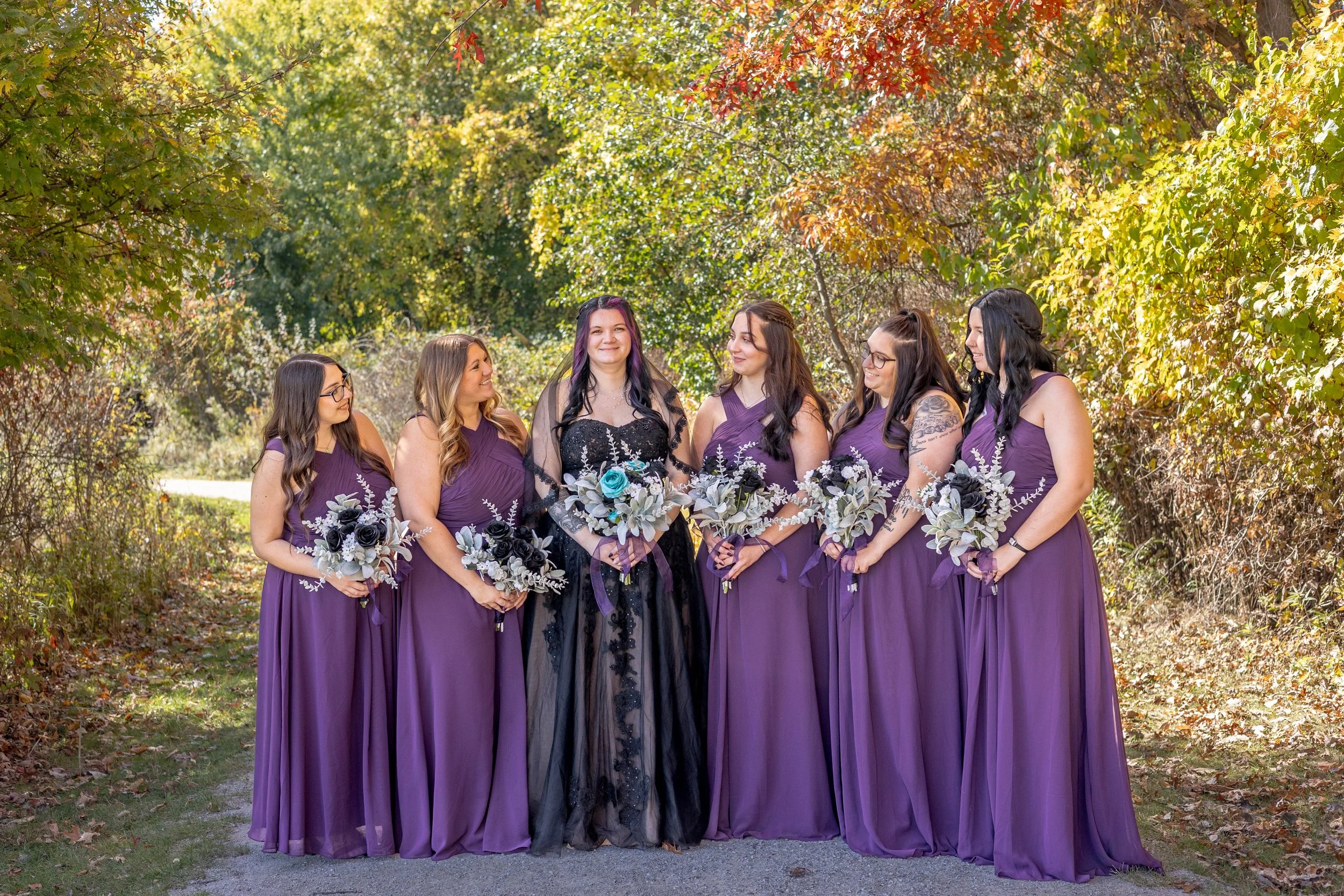 A wedding party of six women, dressed in long purple dresses, stands outdoors in a wooded area with fall foliage. The woman in the center wears black attire with purple hair, holding a bouquet, and is surrounded by bridesmaids holding bouquets, all s