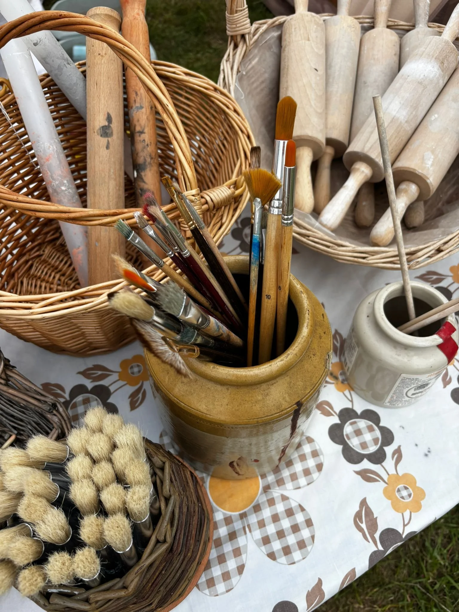 Collection of paintbrushes in ceramic and wicker containers on patterned tablecloth.
