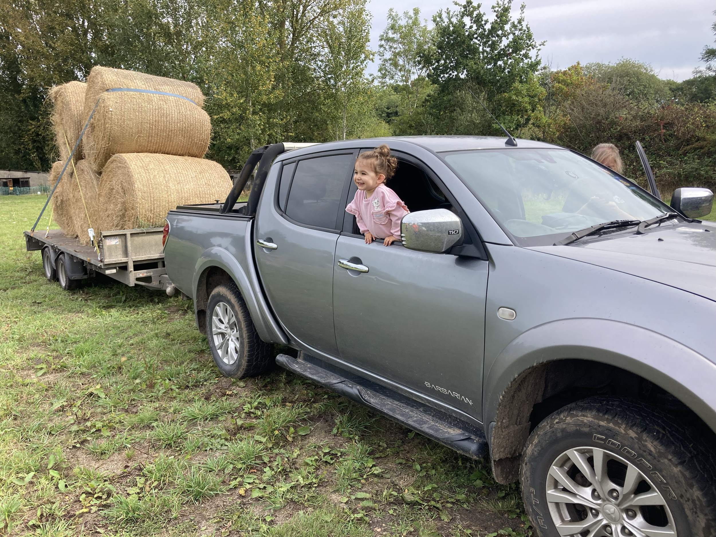 A grey pickup truck with bale hay on a trailer in a grassy field. A girl leaning out of the driver's window, smiling.