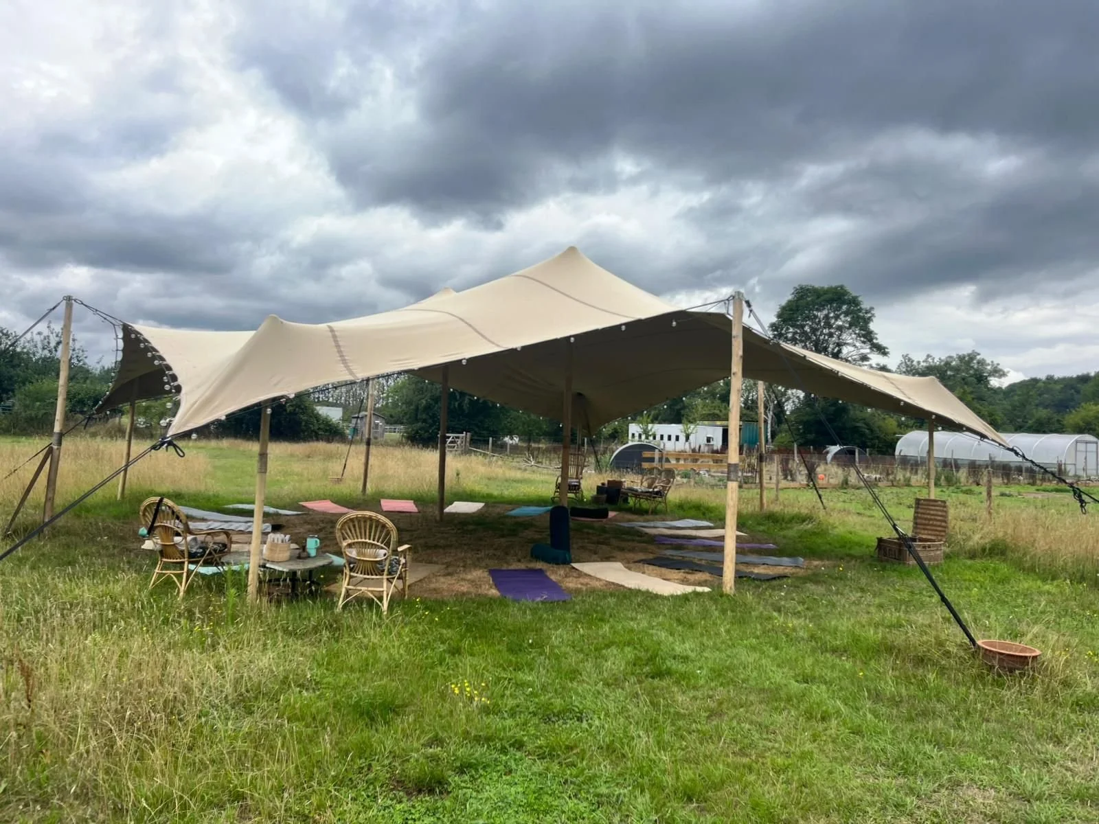 A large outdoor tent with a beige canopy supported by wooden posts, set up in a grassy field. There are colorful mats on the ground, and chairs and a small table with cups and a teapot nearby. The sky is cloudy, and there are farm buildings and trees