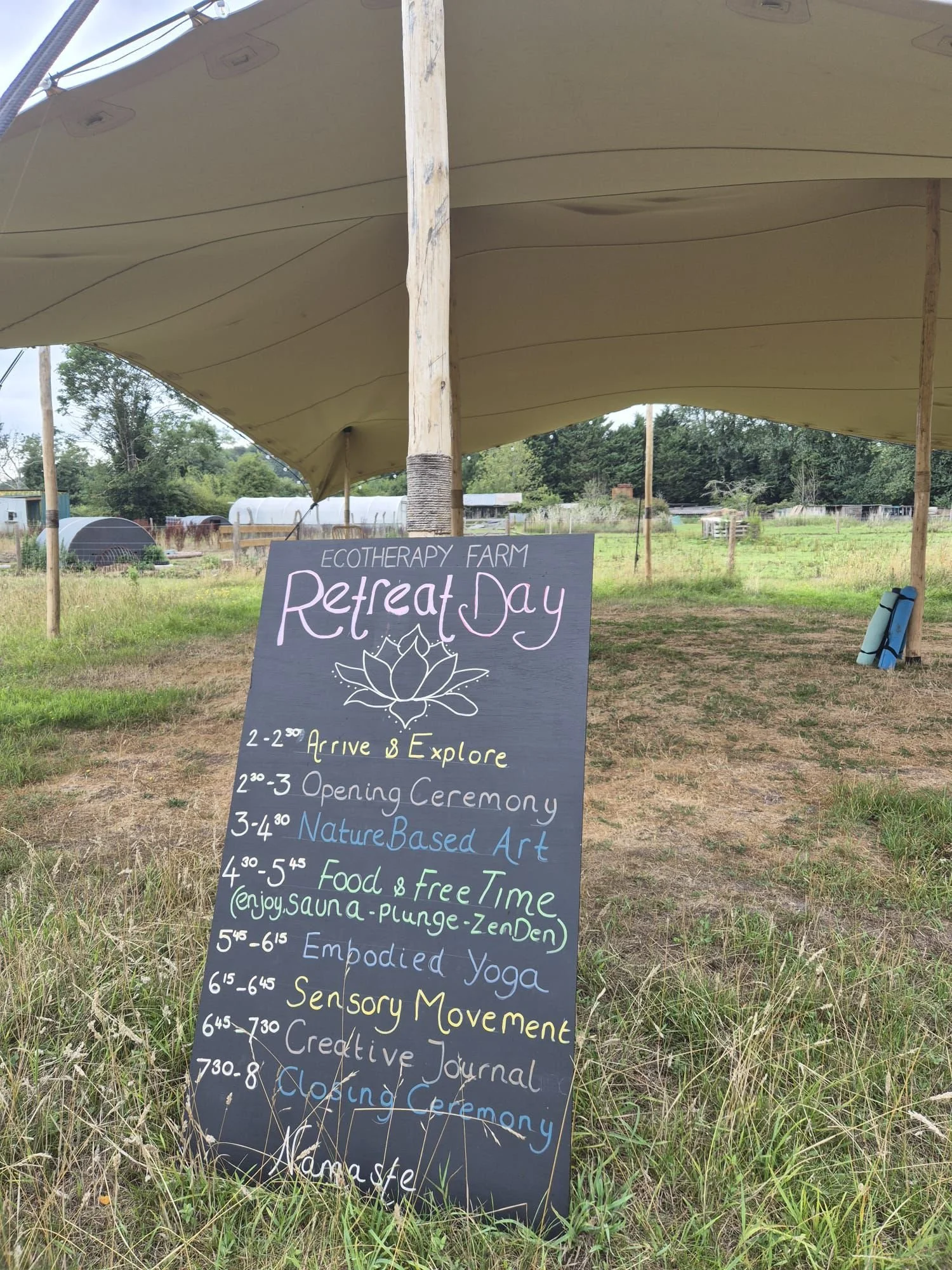 A blackboard sign listing a schedule for Ecotherapy Farm Retreat Day activities under a large tent outdoors.