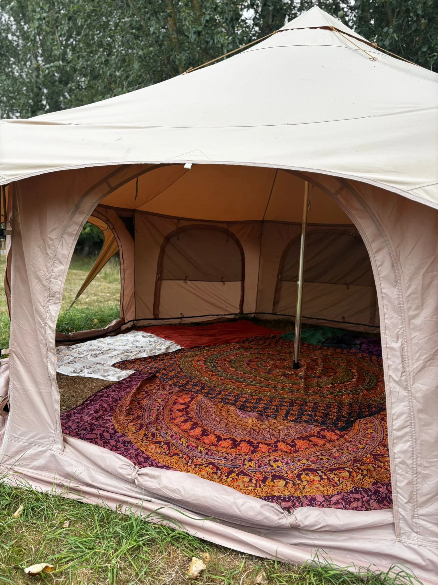 The image shows the interior of a beige camping tent set up outdoors on grass, with Paisley and floral patterned rugs on the ground inside.
