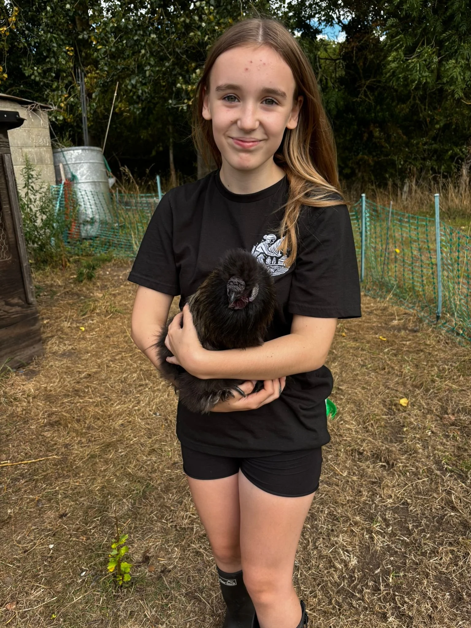 A girl holding a black chicken outdoors in a yard with trees and a green fence.