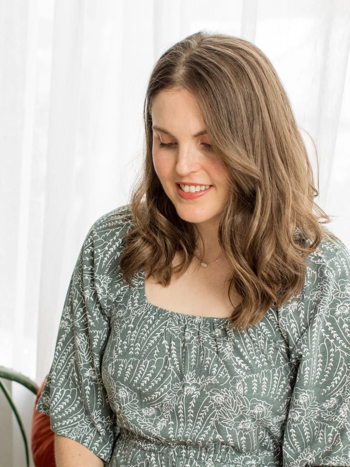 Woman with brown hair in green floral dress, smiling and looking down, sitting indoors.