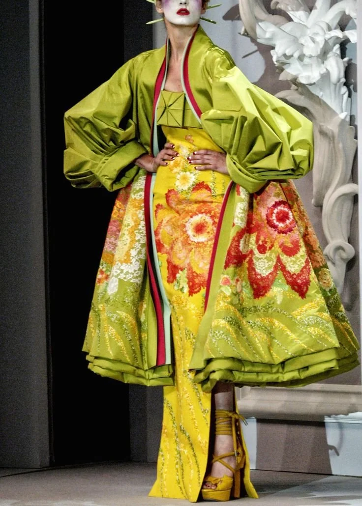 Person dressed in an elaborate yellow floral-patterned gown and matching shoes, standing with hands on hips near a sculpted white floral art piece.