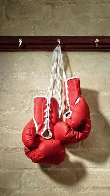 A pair of red boxing gloves hanging from white shoelaces tied to a wooden hook on a beige brick wall.