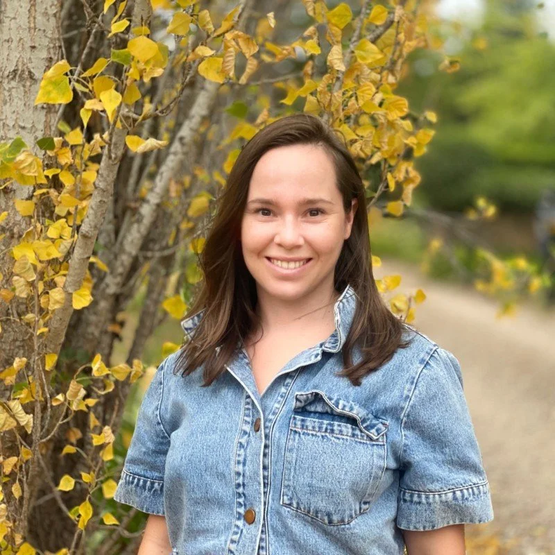 A young woman with shoulder-length brown hair, smiling, wearing a light blue denim jacket, standing outdoors next to a tree with yellow leaves.