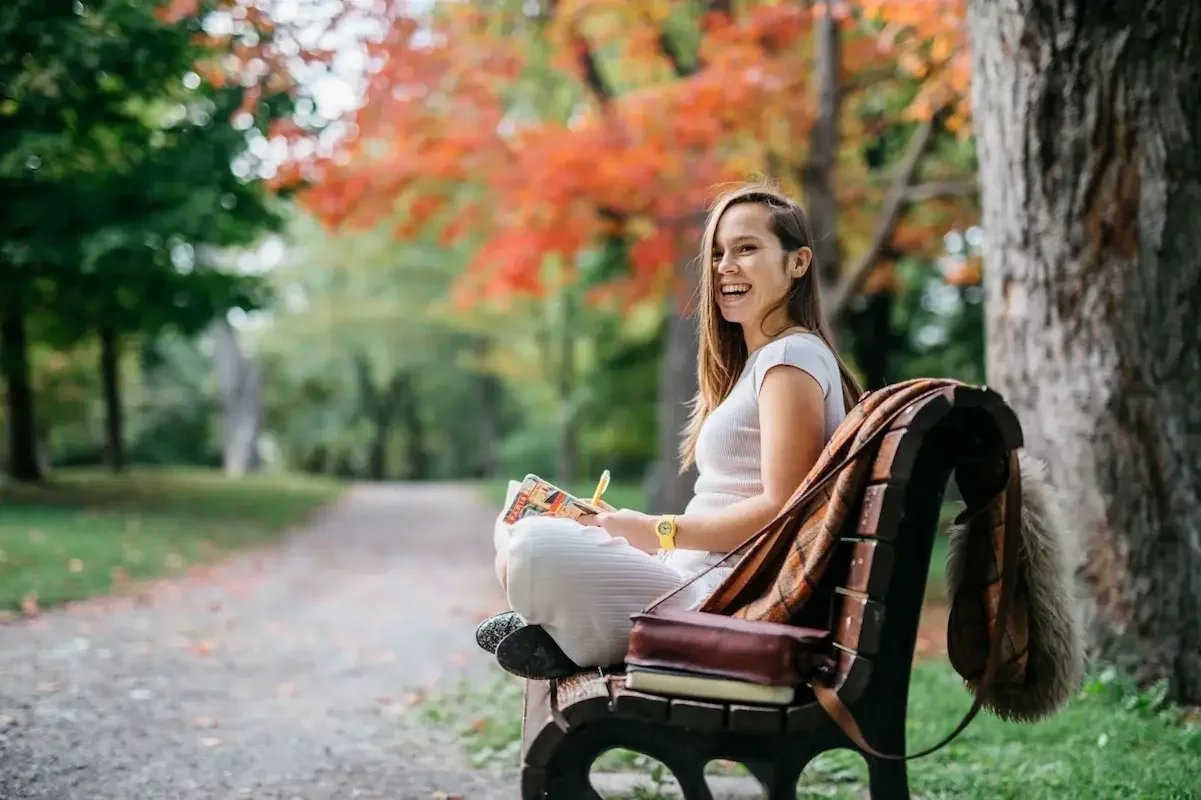 A young woman sitting on a park bench with a large tree and autumn-colored leaves behind her. She is smiling, holding a book and a pencil, with a backpack hanging on the side. The scene is outdoors on a gravel path surrounded by green and orange foliage.