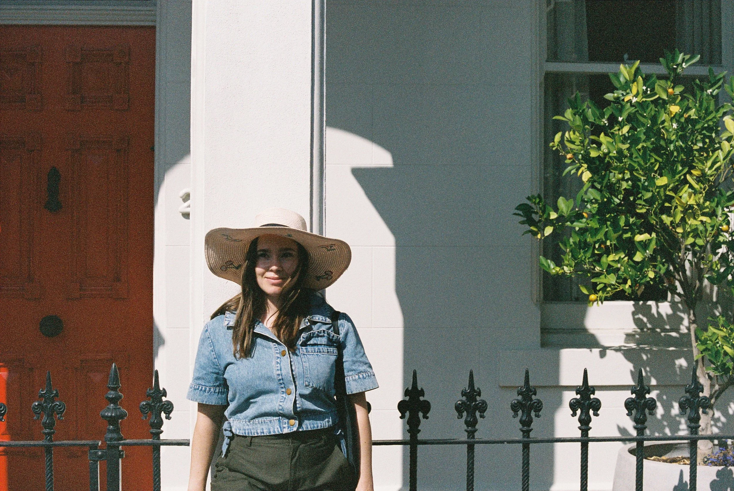 A woman in a denim shirt and wide-brimmed straw hat standing in front of a white house with a red door, black iron fence, and green shrubbery, illuminated by sunlight.