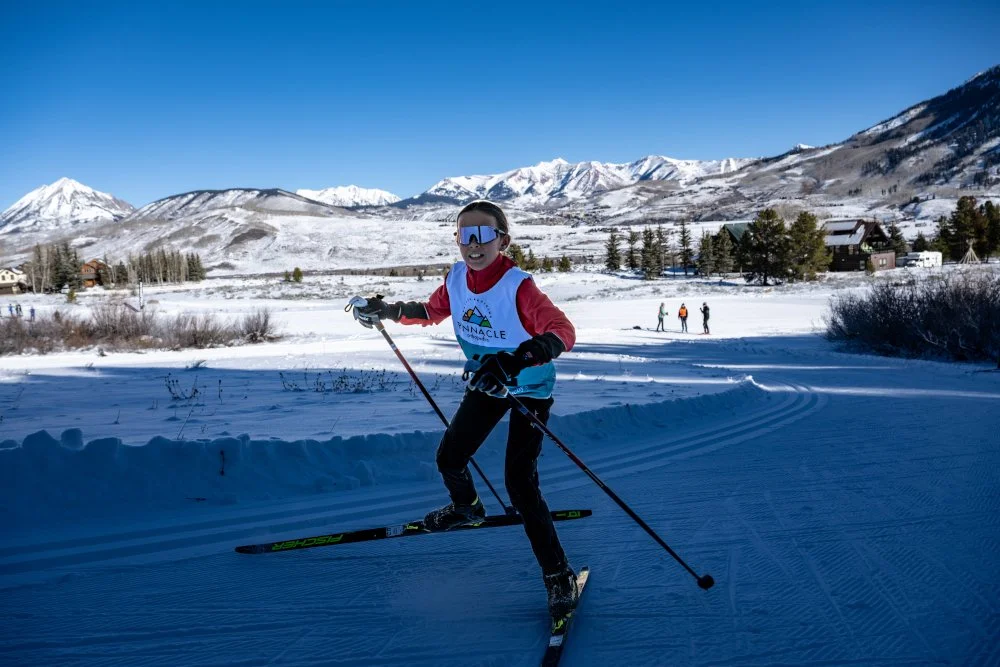A young racer skis through the shadows with mountains and a blue sky behind them