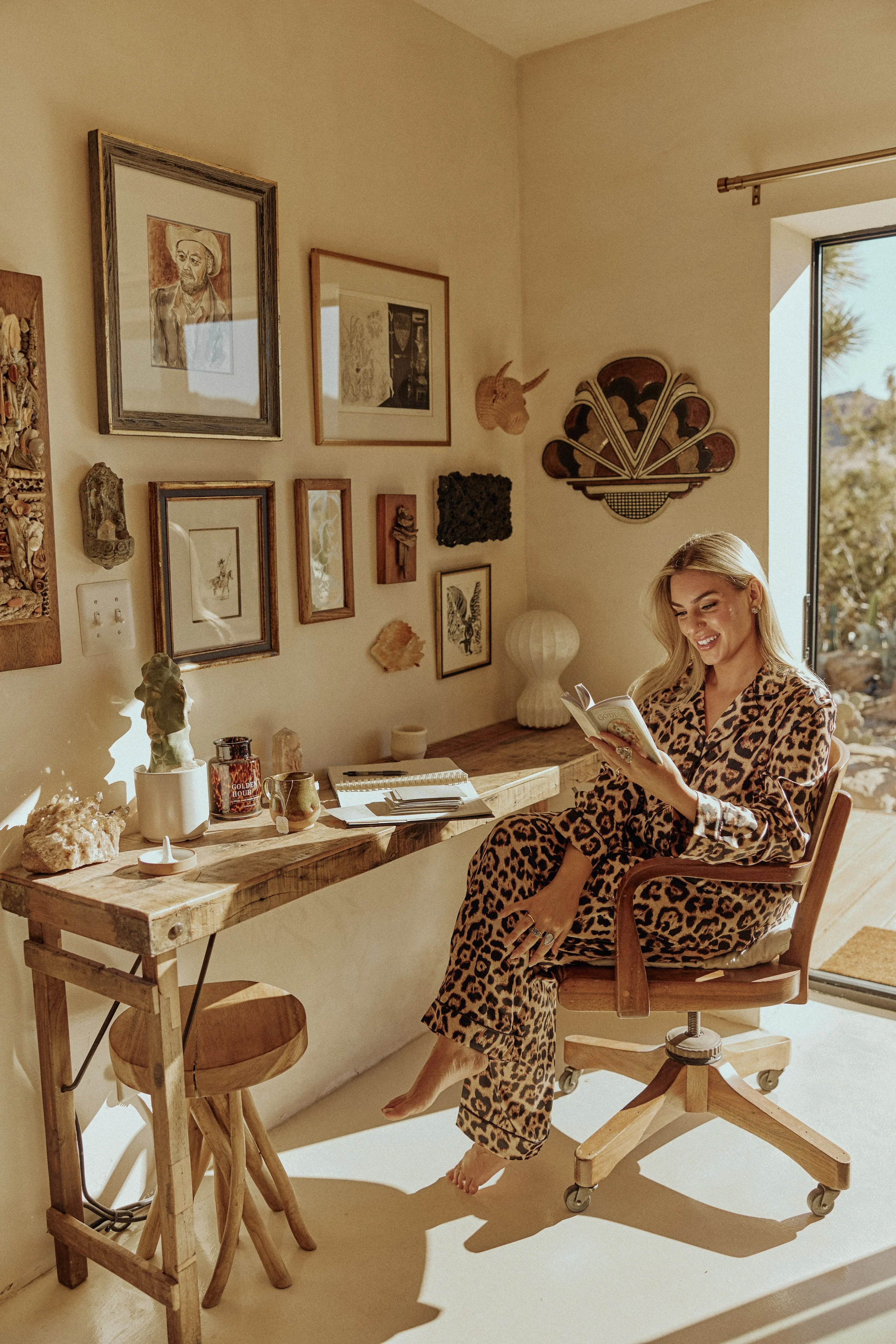 journaling at a wooden desk in a sunlit Joshua Tree home, wearing leopard-print loungewear with crystals and artwork