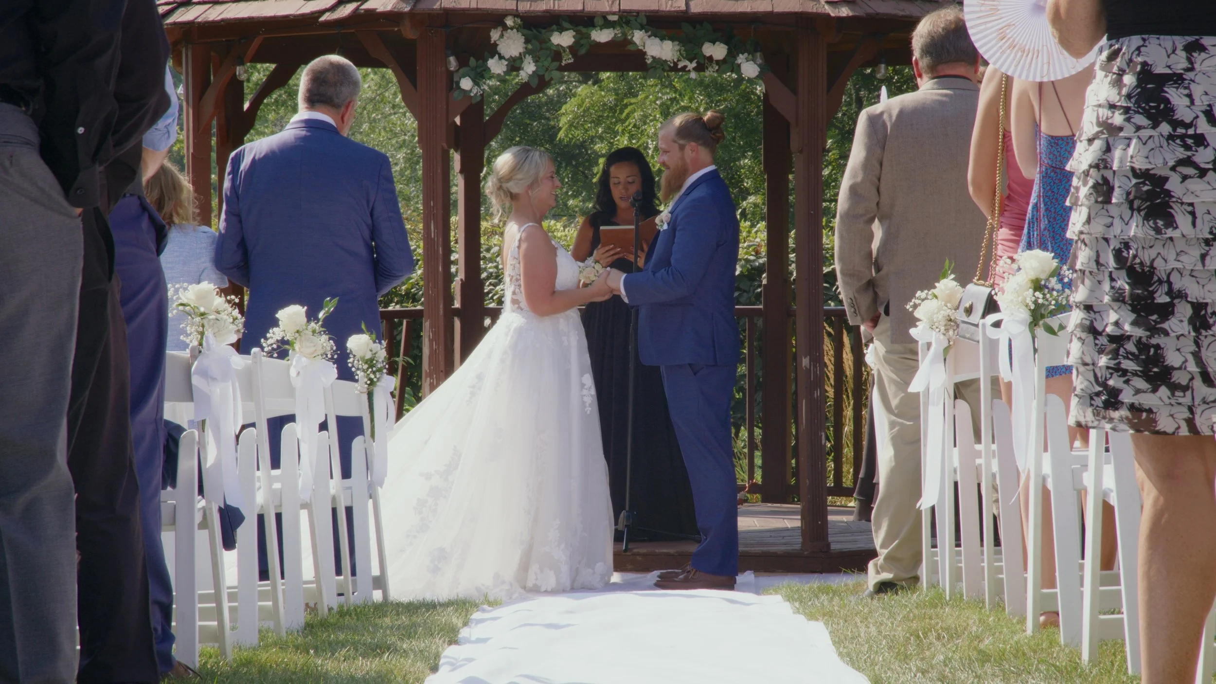 A couple exchanging wedding vows under a wooden gazebo decorated with white flowers, with an officiant reading from a book, surrounded by guests dressed in formal attire.