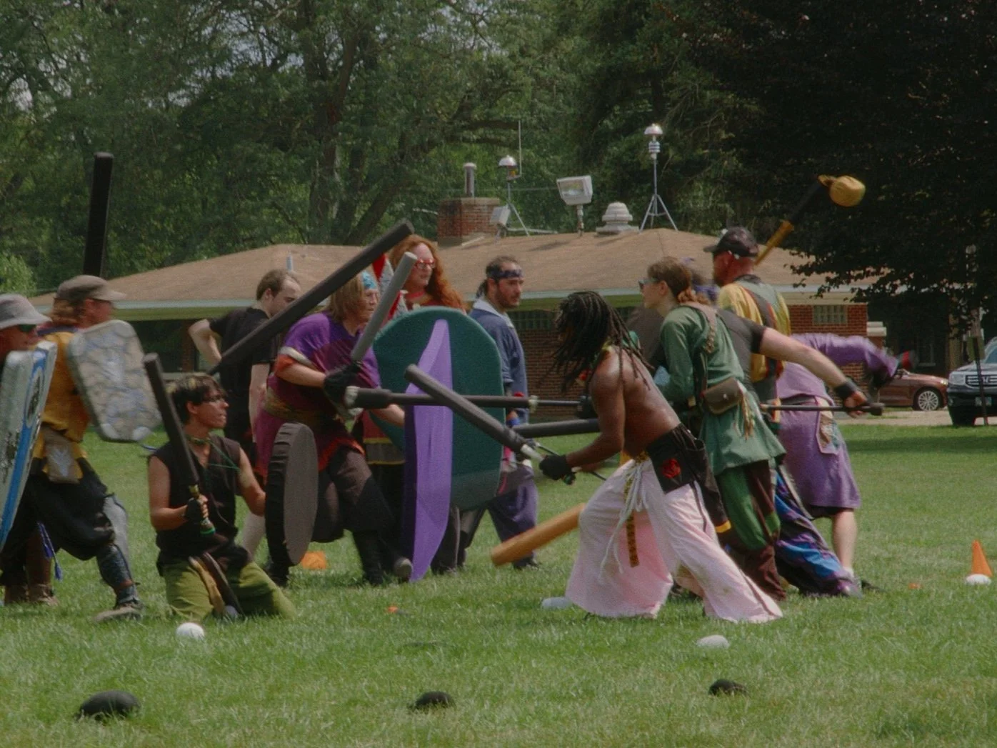 Group of people engaging in a mock medieval battle with foam weapons, shields, and costumes on a grassy field.