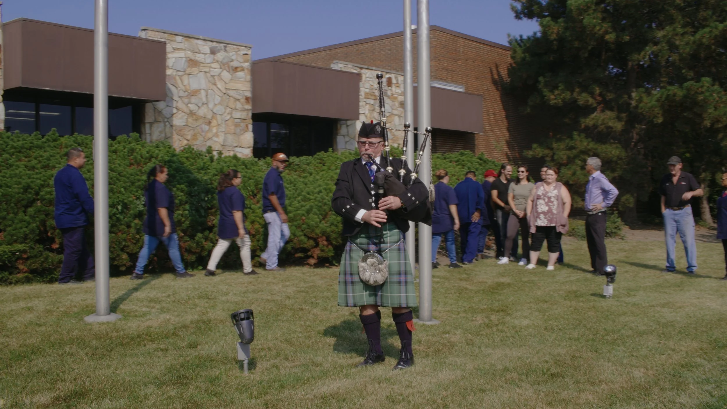 A man dressed in traditional Scottish kilt and playing bagpipes standing on a grassy lawn, with a group of people and a stone and brick building in the background.