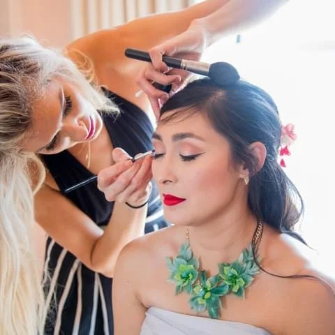 Woman with black hair and floral necklace getting makeup applied by a makeup artist.