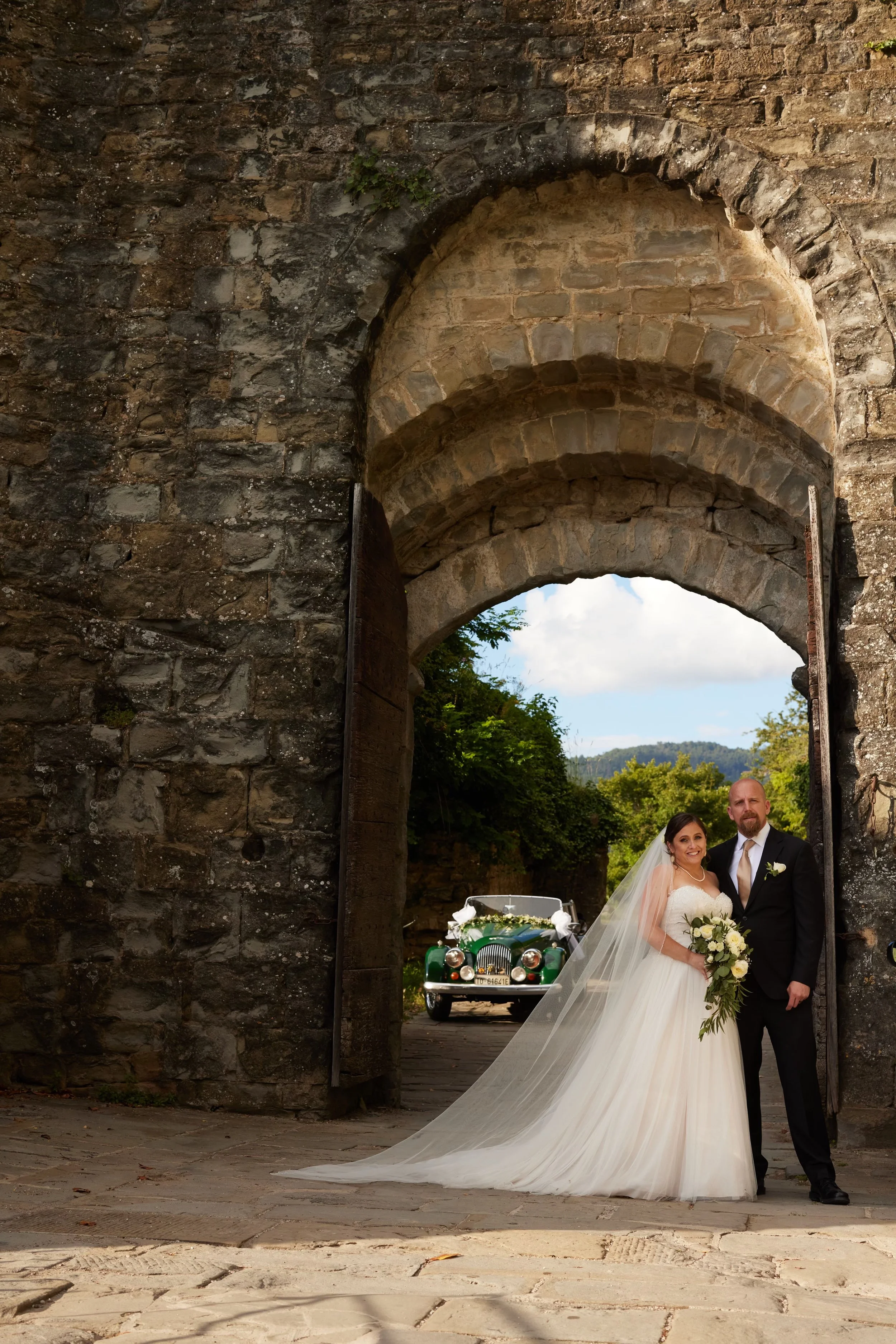 A bride and groom standing together in front of an old stone archway, with a vintage green car decorated with flowers in the background.