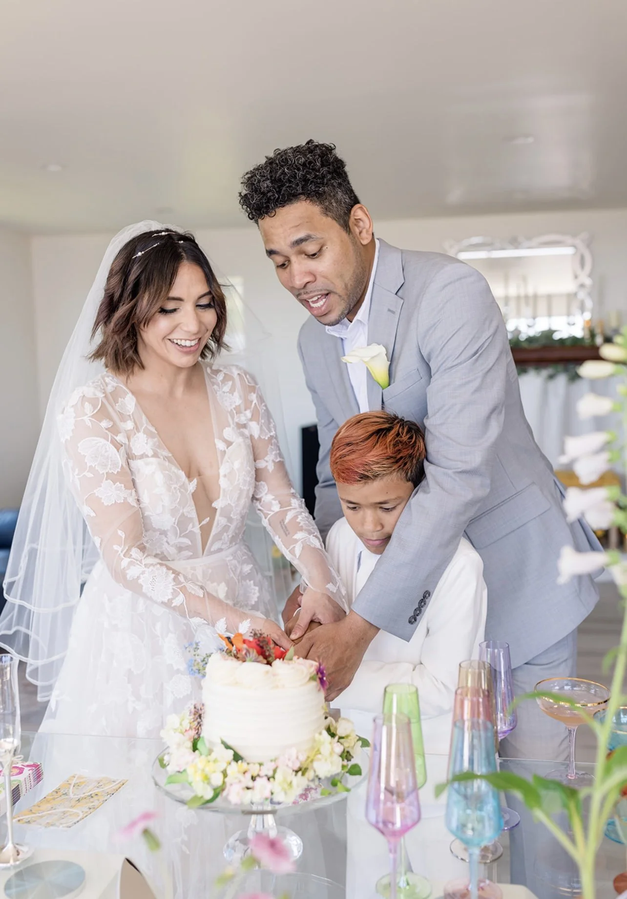 A bride and groom with a young boy, celebrating a wedding with a cake. The bride is wearing a lace wedding dress and veil. The groom is in a grey suit with a white flower boutonniere. They are smiling and cutting the cake together at a decorated tabl