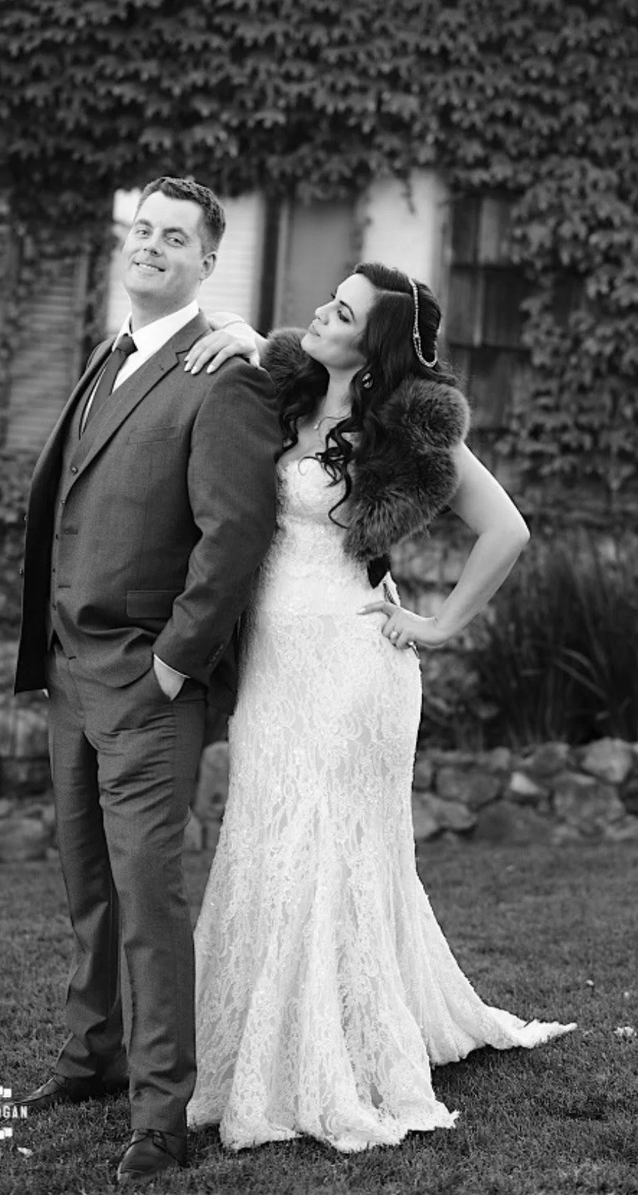 A man in a suit and a woman in a lace wedding dress with a fur stole posing outdoors.