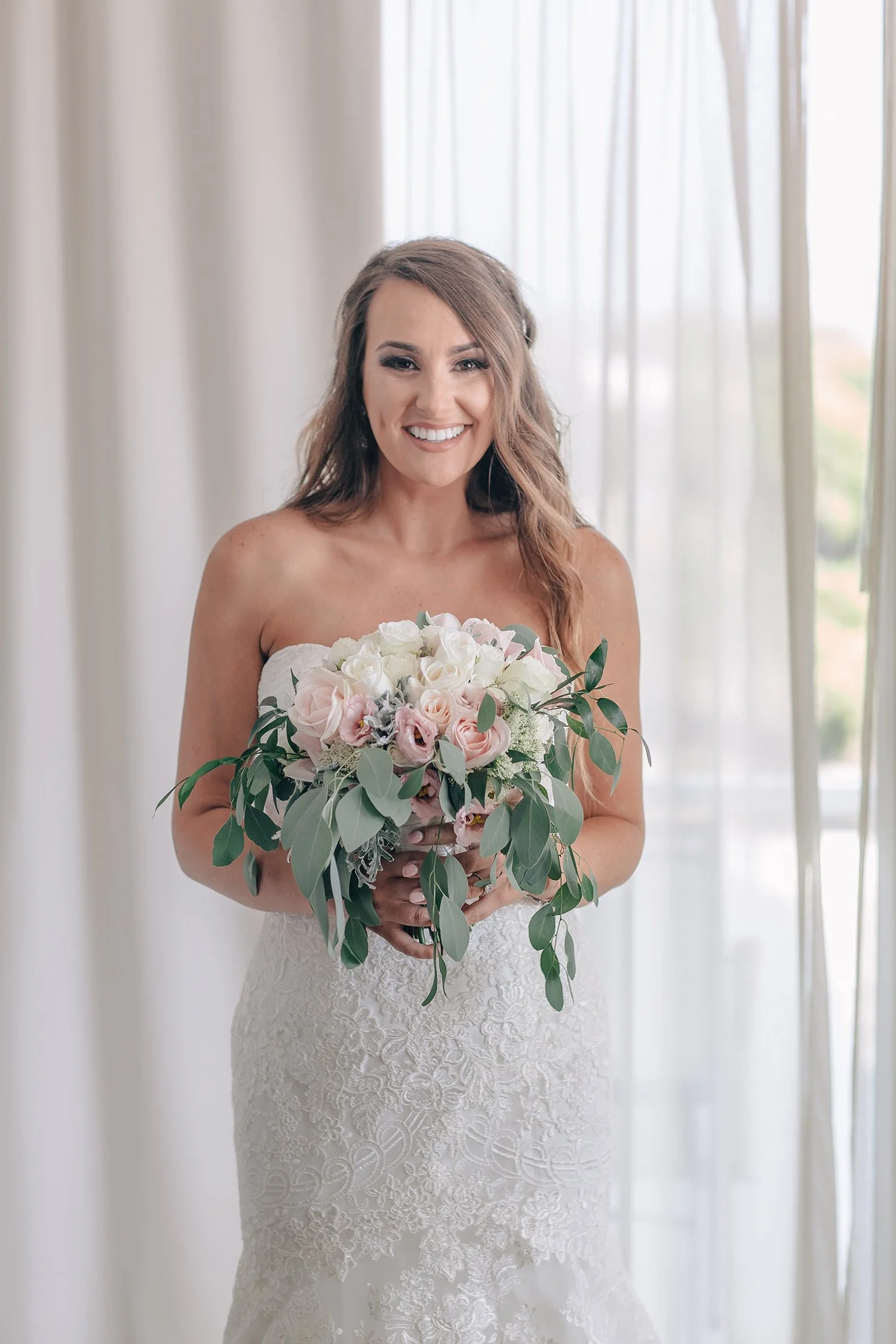 A bride in a strapless lace wedding gown holding a bouquet of pink and white roses with green leaves, smiling in front of sheer curtains.