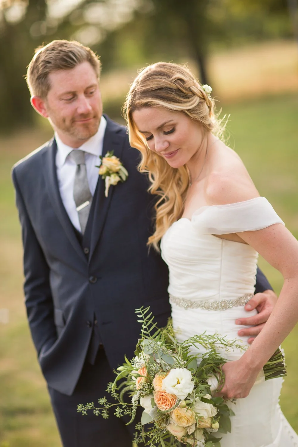 A bride and groom standing outdoors, with the bride holding a bouquet of flowers, both smiling softly. The groom is wearing a dark suit with a boutonniere, and the bride is in a white dress with a bejeweled belt and a floral hairpiece, surrounded by 