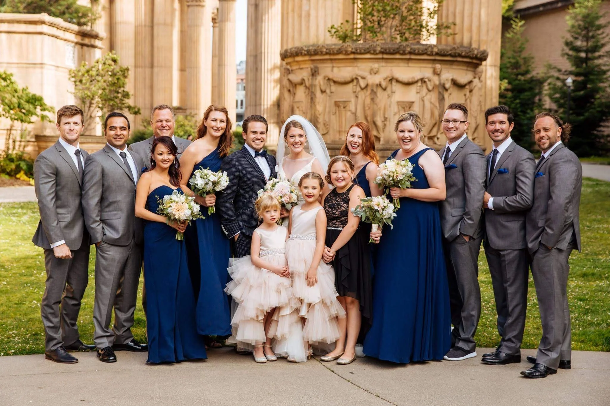 Group of wedding party members, including bride, groom, bridesmaids, groomsmen, and flower girls, posed outdoors in front of a historic architectural structure, all smiling at the camera.