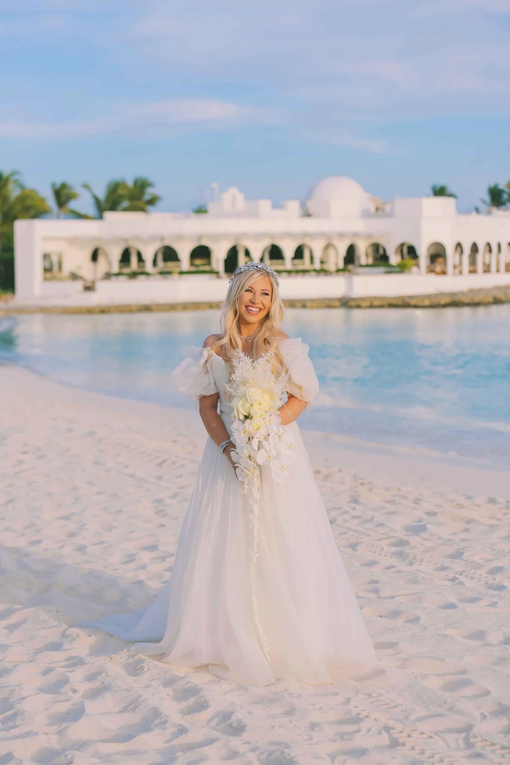 A smiling bride in a white wedding dress holding a bouquet of white flowers standing on a sandy beach with a white building and palm trees in the background.