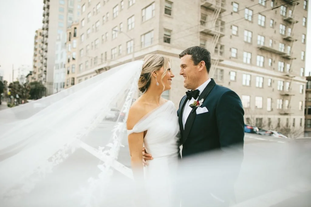 Bride and groom looking at each other, standing close with urban buildings in the background, the bride wearing a white wedding dress and veil, the groom in a dark tuxedo and bow tie.