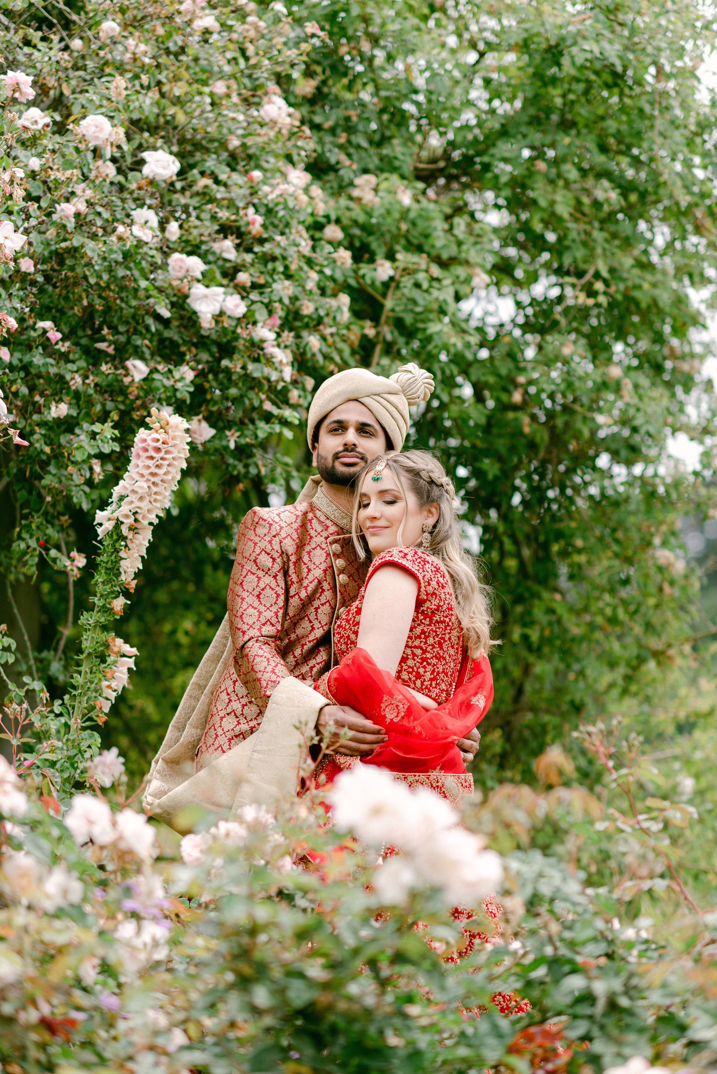 A couple in traditional Indian wedding attire poses among flowering bushes. The groom wears a beige turban and a patterned golden-red sherwani, while the bride wears a red ornate saree with gold embroidery and jewelry, standing with eyes closed, surr