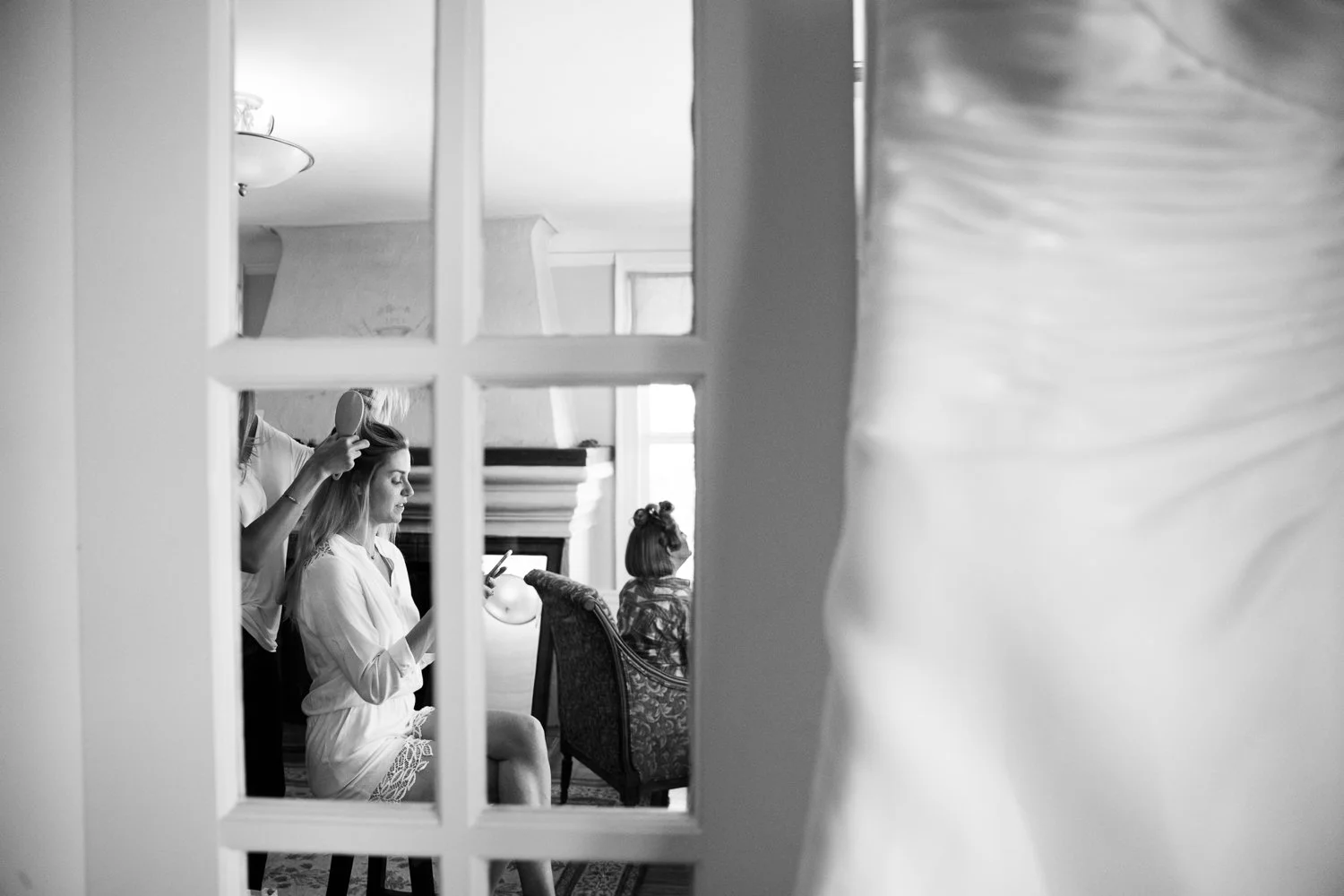 A woman getting her hair styled while sitting on a chair, with another woman looking at her phone, and a young girl sitting in a chair, as seen through a glass window.