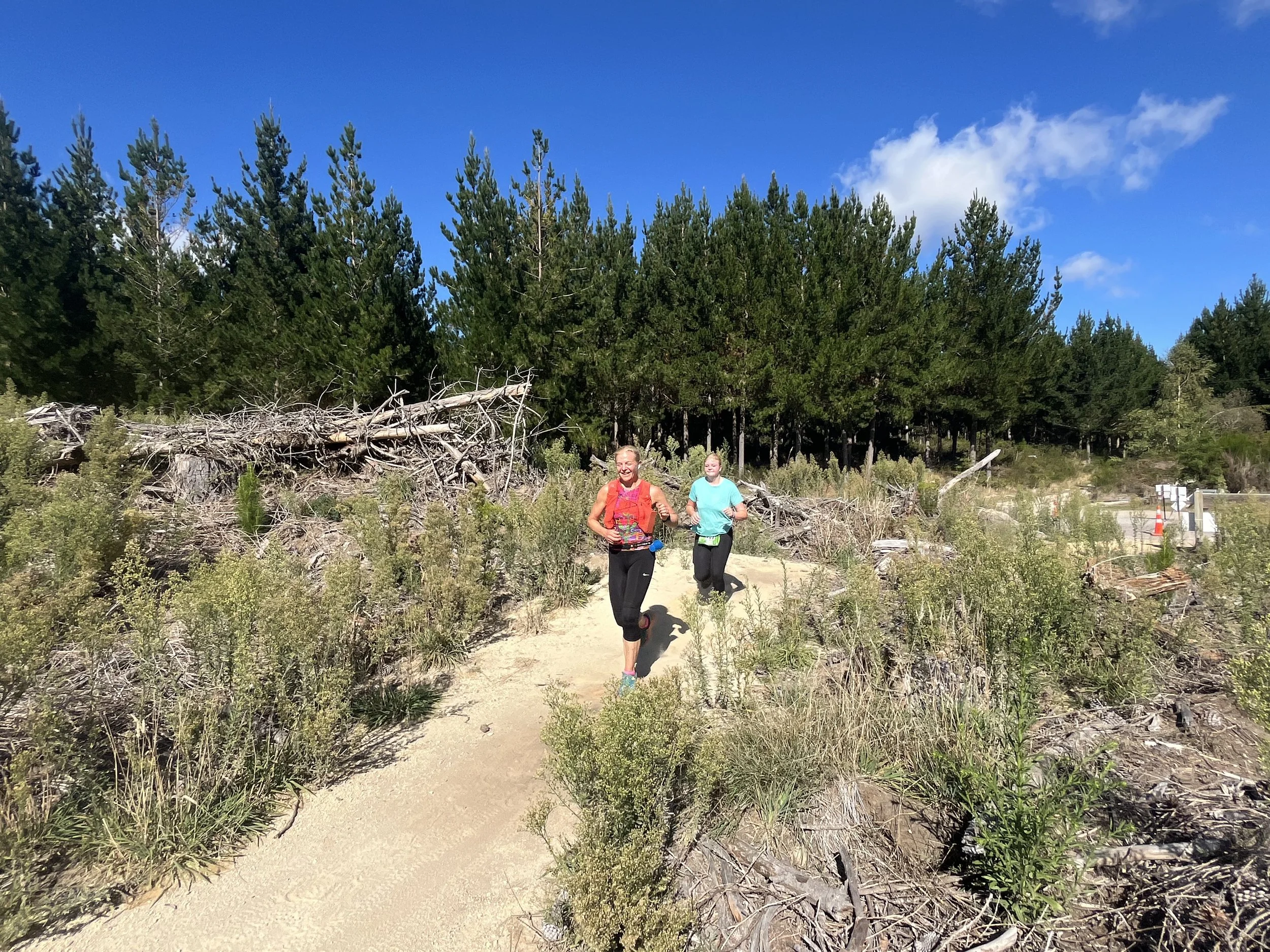 Two women running on a trail through a forested area with trees and fallen logs under a blue sky.