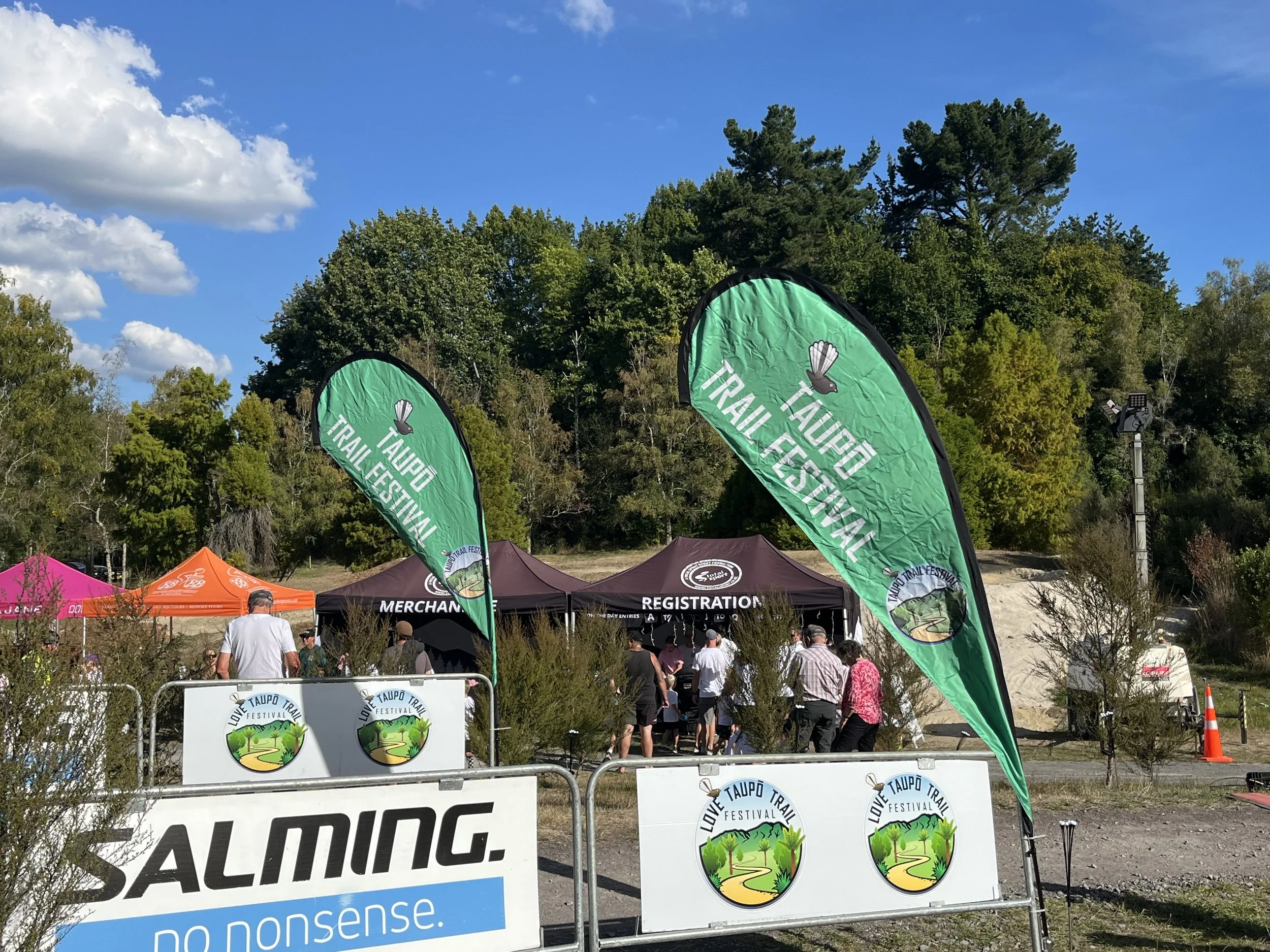 People attending the Taupo Trail Festival, with registration tents and green flags, outdoors in a park with trees and a blue sky.