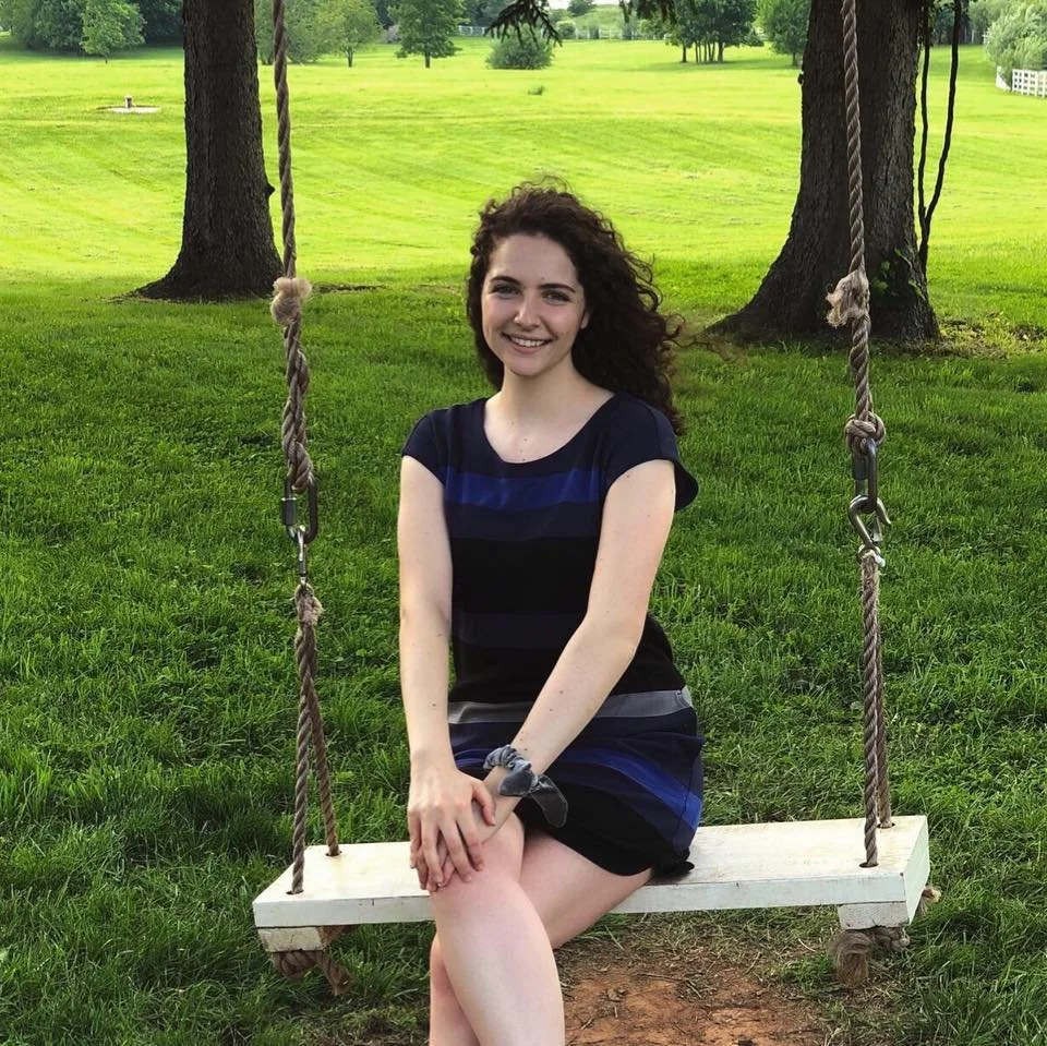 A young woman with curly brown hair, smiling, seated on a wooden swing hanging from two trees in a grassy park.