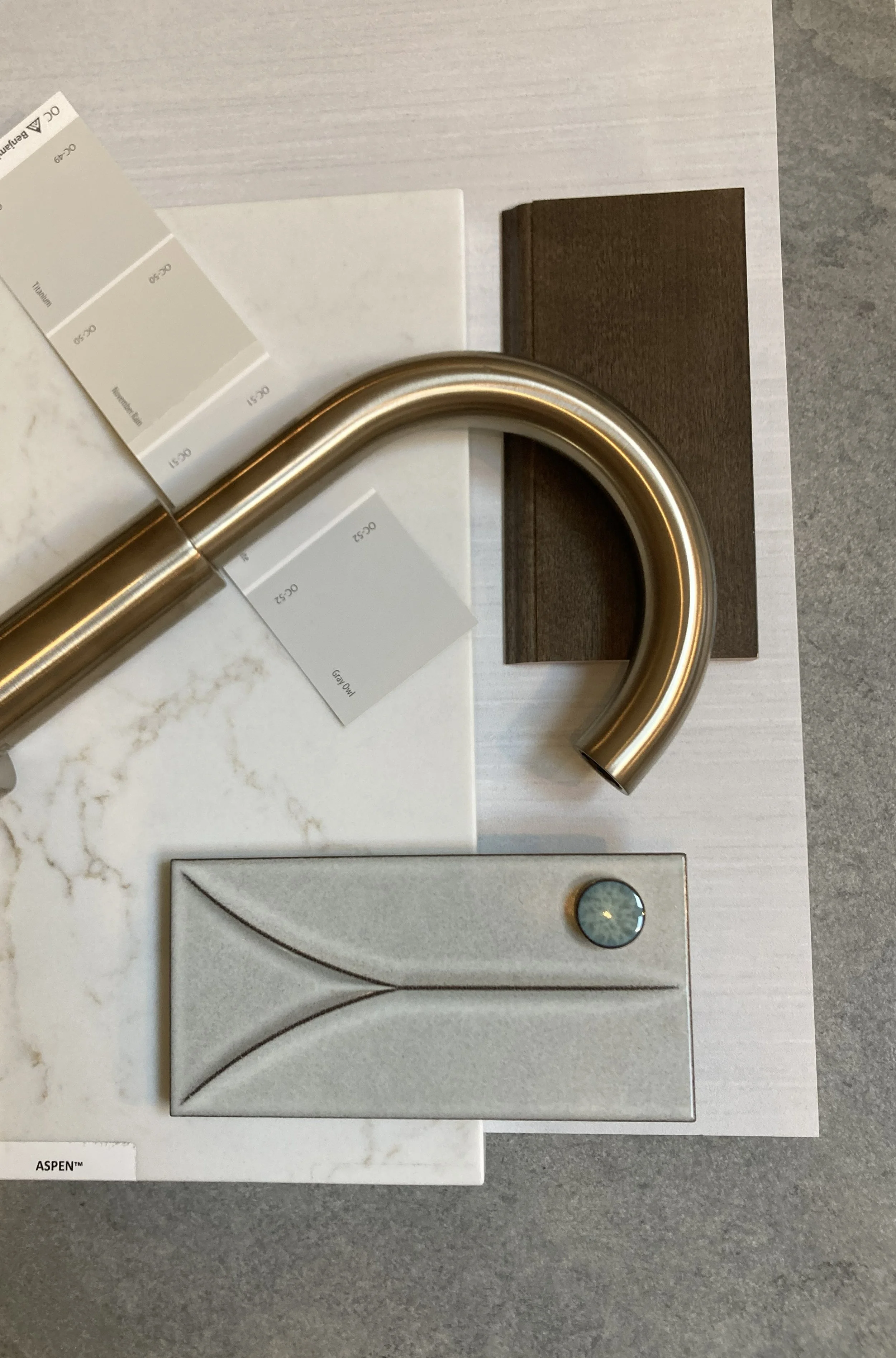 A collection of interior design materials, including a brushed gold kitchen faucet, a white marble tile sample, a dark wood sample, a grey textured tile with a circular decorative element, and color samples, all arranged on a light-colored surface.