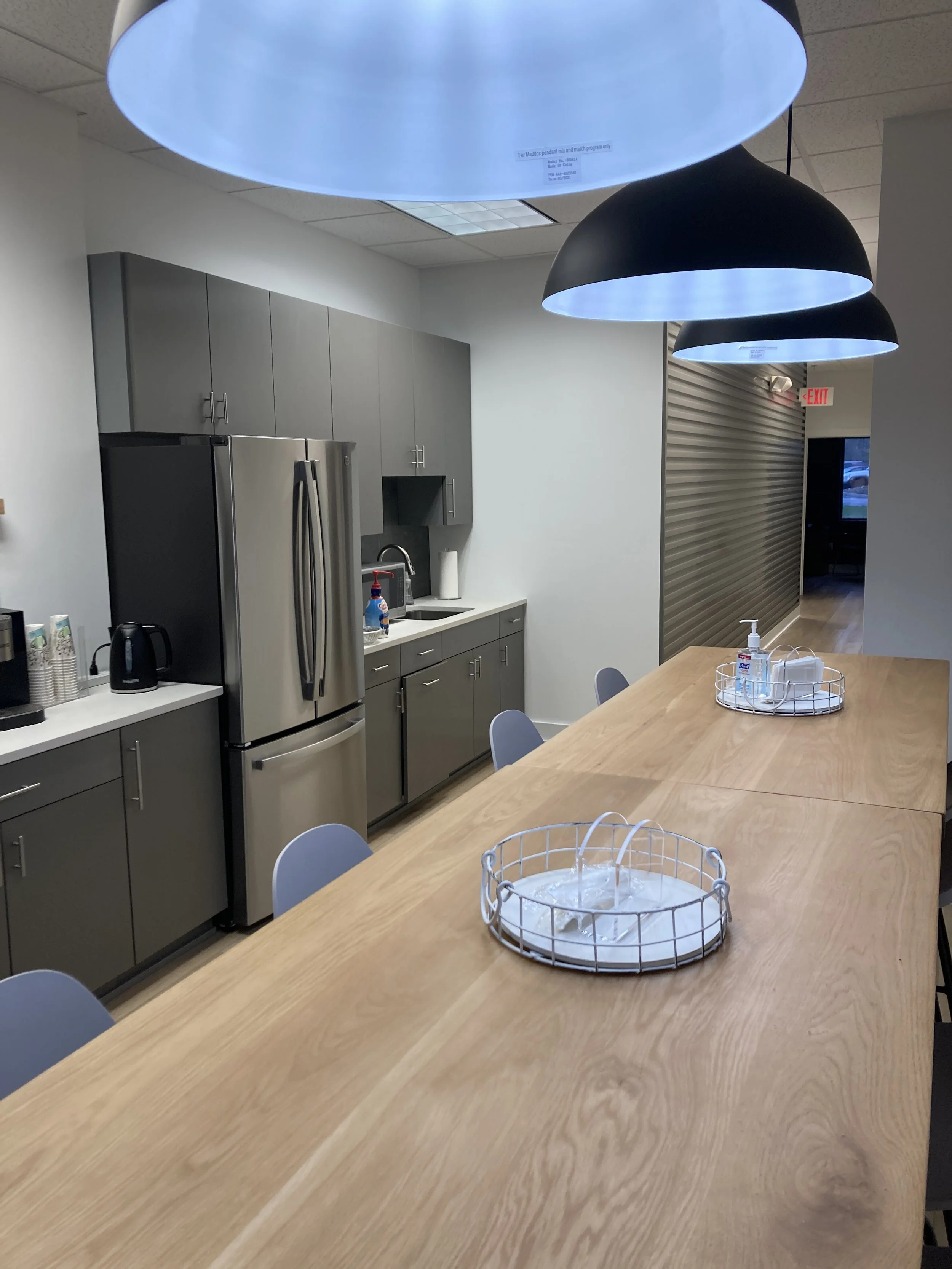 A modern kitchen or communal area with a long wooden table, gray cabinets, stainless steel refrigerator, black pendant lights, hand sanitizer, and chairs.