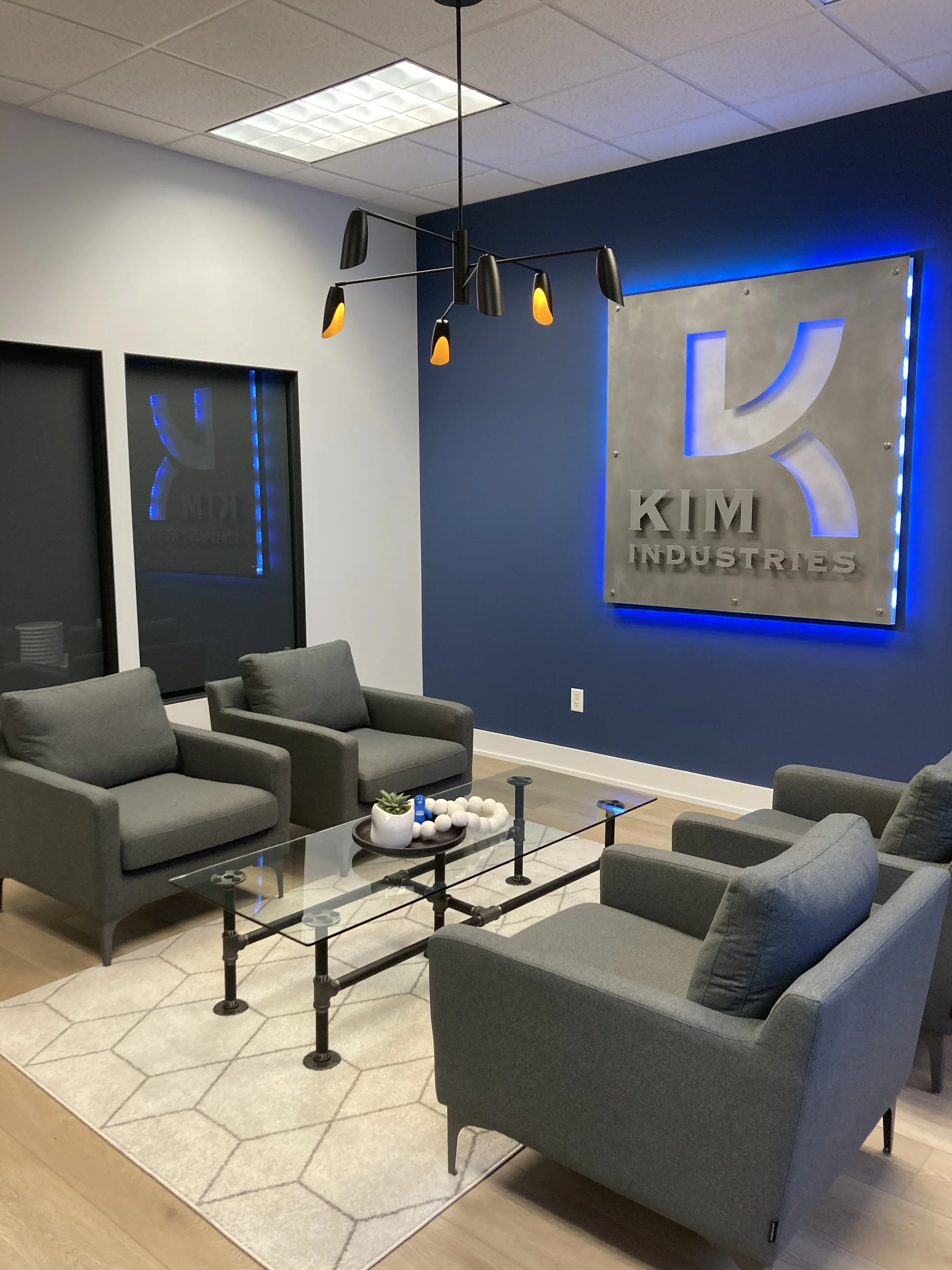 Modern office lounge with gray armchairs arranged around a glass coffee table. The wall displays a large illuminated logo for 'Kim Industries' with a blue backlight. A contemporary black chandelier with orange accents hangs from the ceiling, and a beige rug with a geometric pattern is beneath the seating area.