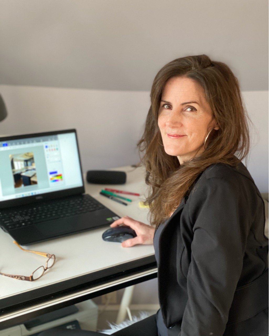 A woman with brown hair and hoop earrings sitting at a desk using a laptop, with a computer mouse in hand, in a well-lit room.