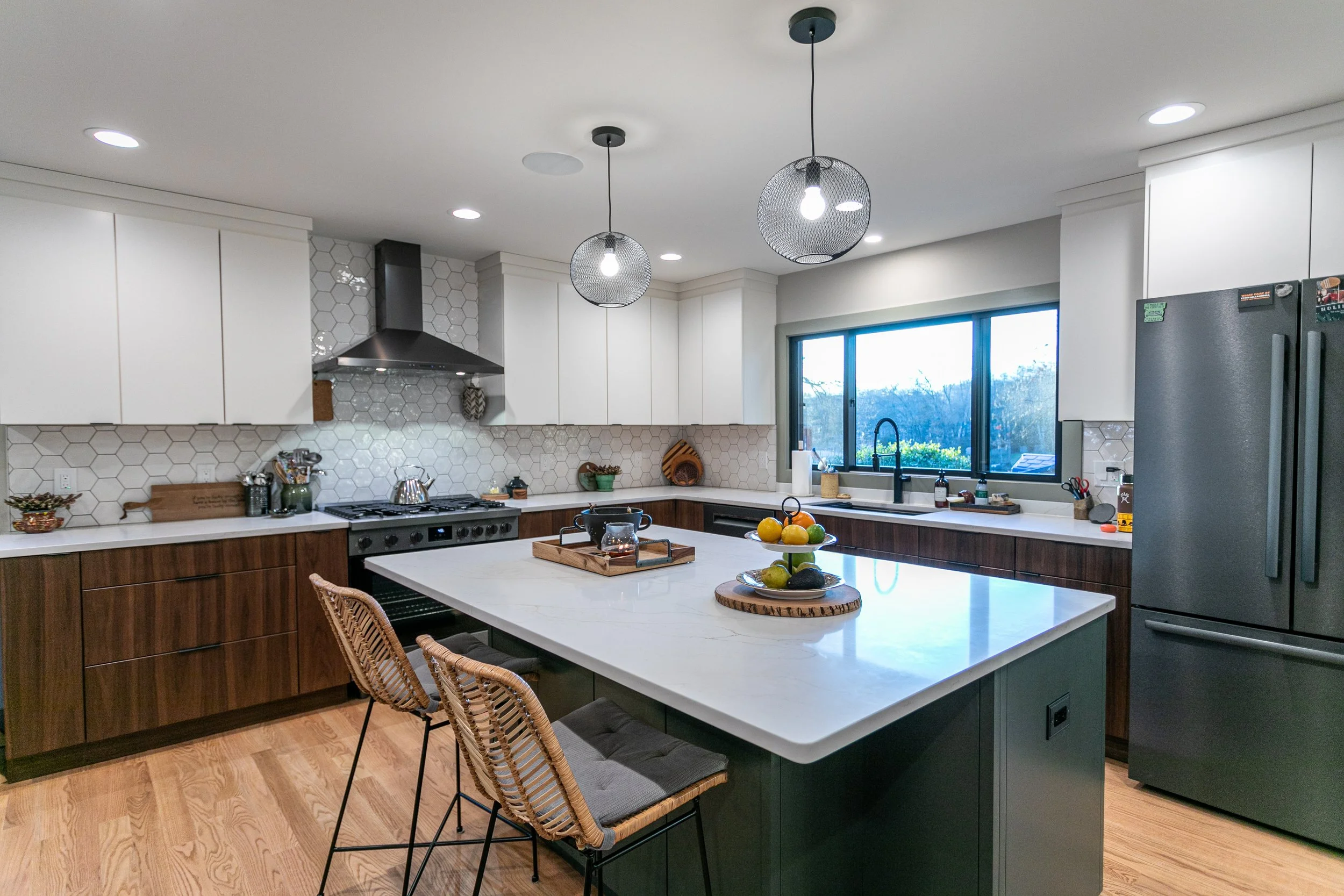 Modern kitchen with white cabinets, dark wooden lower cabinets, and a large white island with a marble top. There are two rattan chairs with cushions at the island. The kitchen has a black refrigerator, a stainless steel range hood, and a window above the sink. The backsplash features hexagonal tiles. Two pendant lights hang over the island, and the floor is wooden.