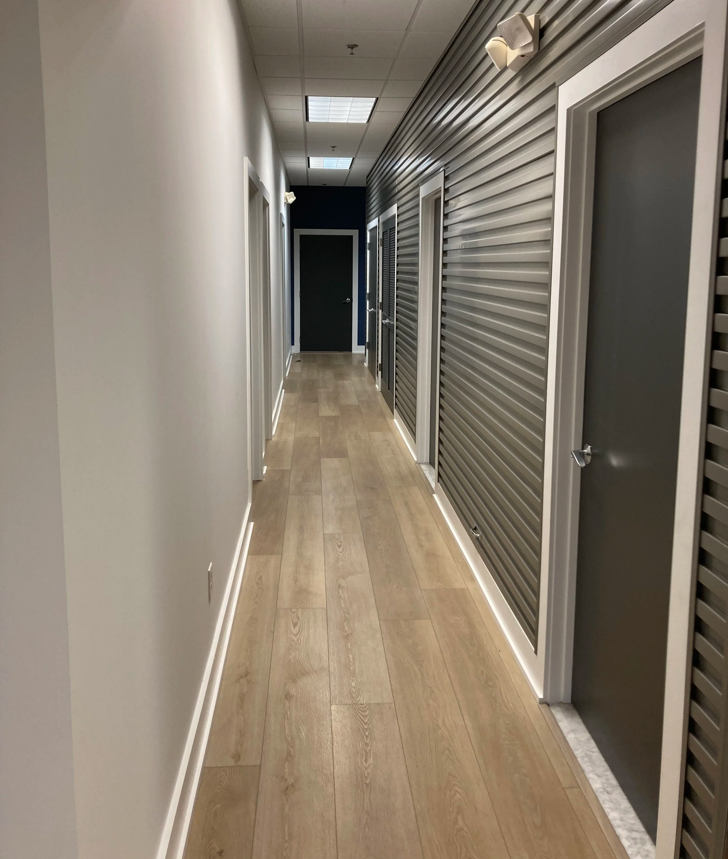Empty hallway in an office building with a light wood floor, white wall on the left, and gray metal doors with vents on the right. Ceiling tiles with fluorescent lights and a dark door at the end of the hall.