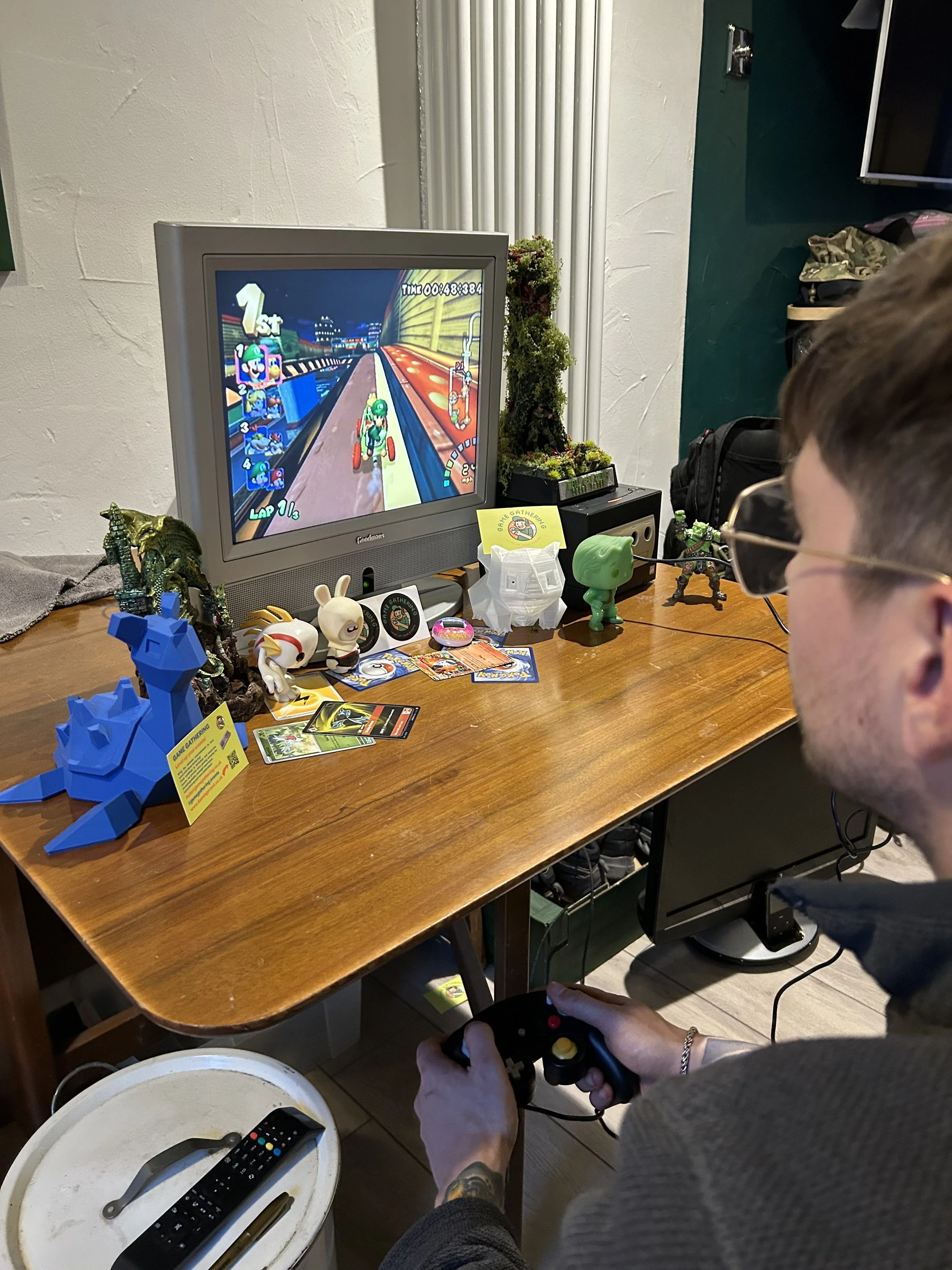 A man playing a racing video game on a monitor, holding a game controller, with various action figures and collectibles on the table.