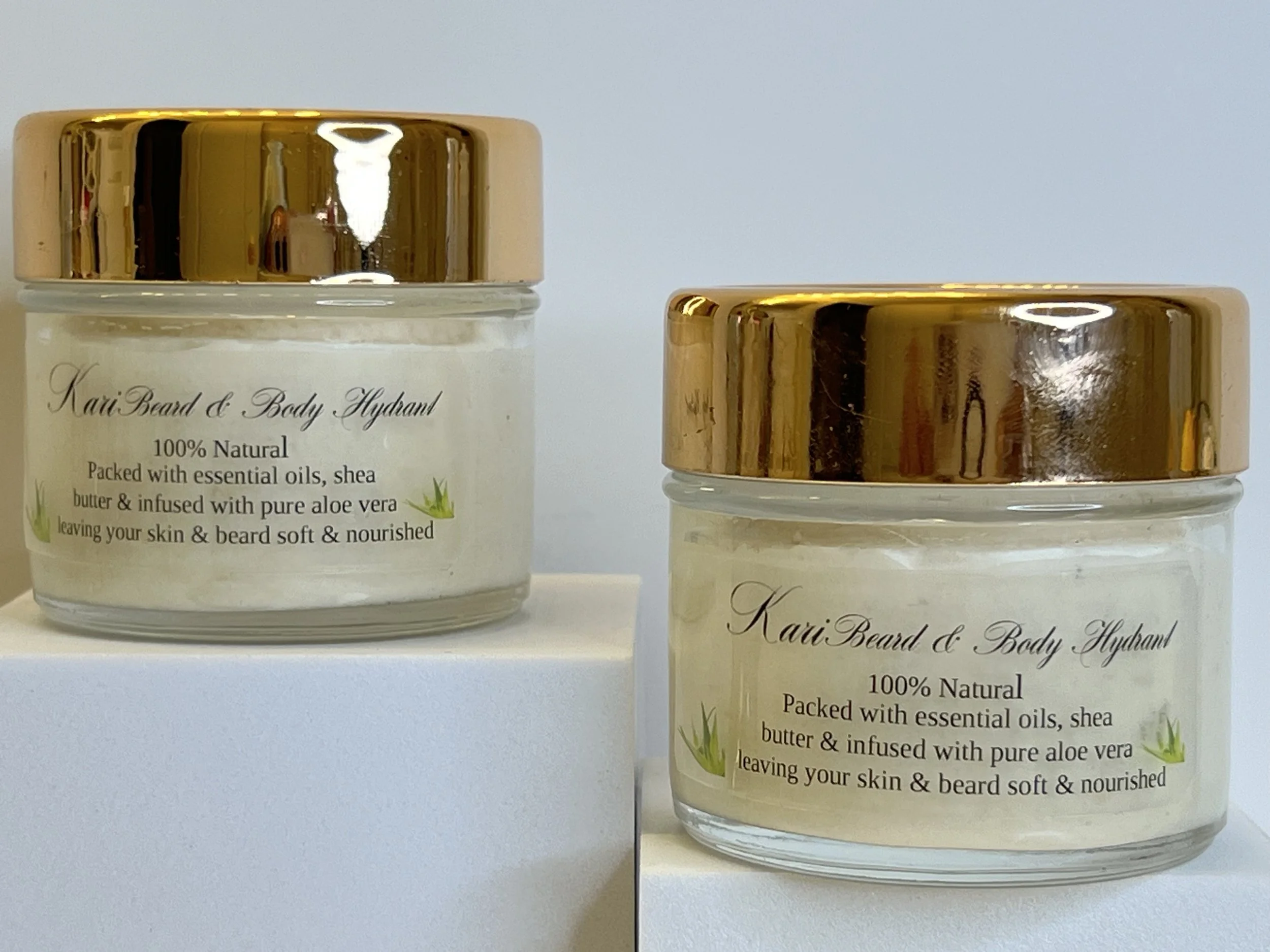 Two jars of Kari Beard & Body Hydrant cream with gold lids, clear glass containers, and labels indicating natural ingredients like essential oils, shea butter, and aloe vera.