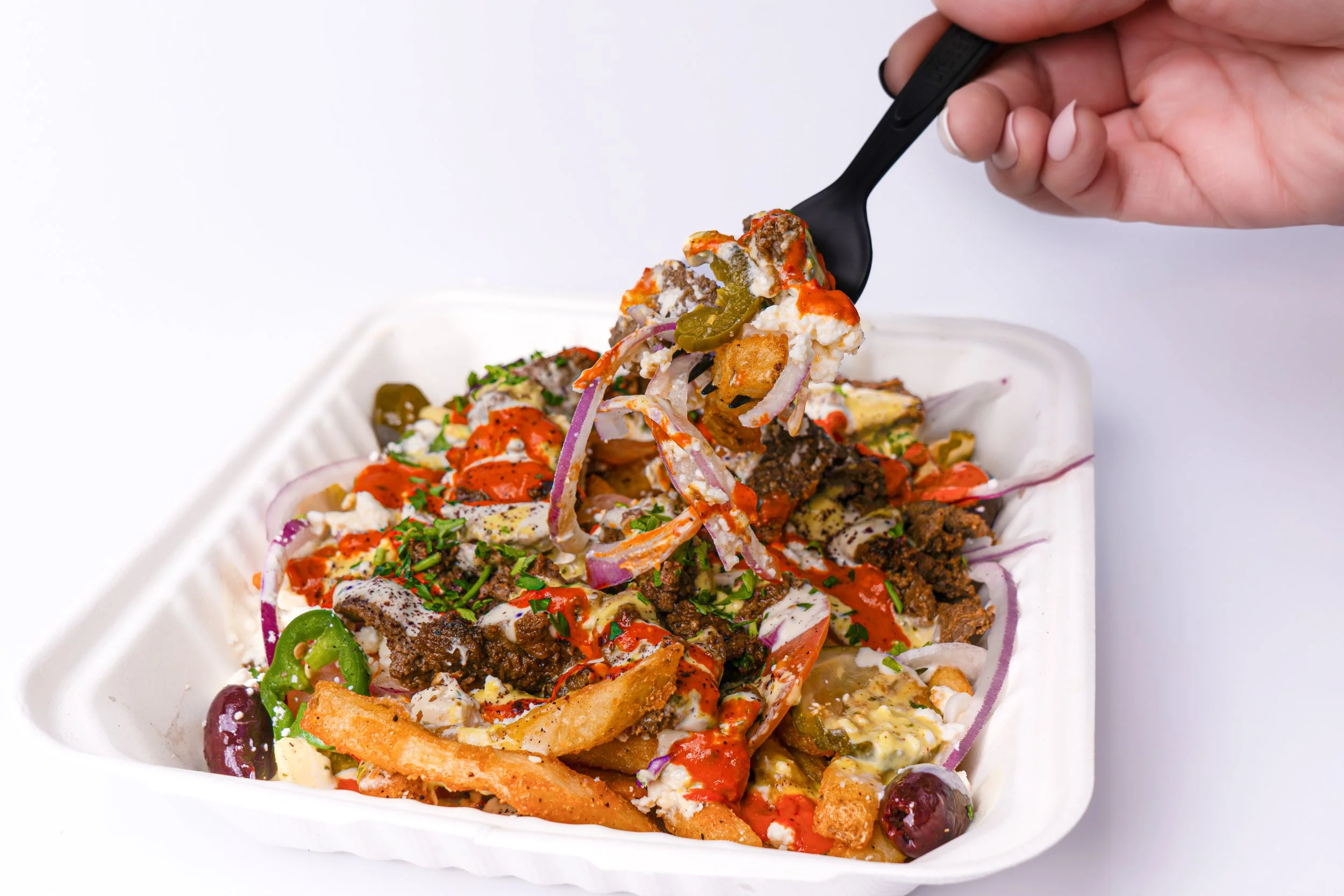 Person holding a black fork with a bite of loaded fries topped with cheese, chili, onions, and sauce, placed over a white styrofoam container filled with loaded fries, chili, onions, jalapeños, and garnished with chopped herbs.