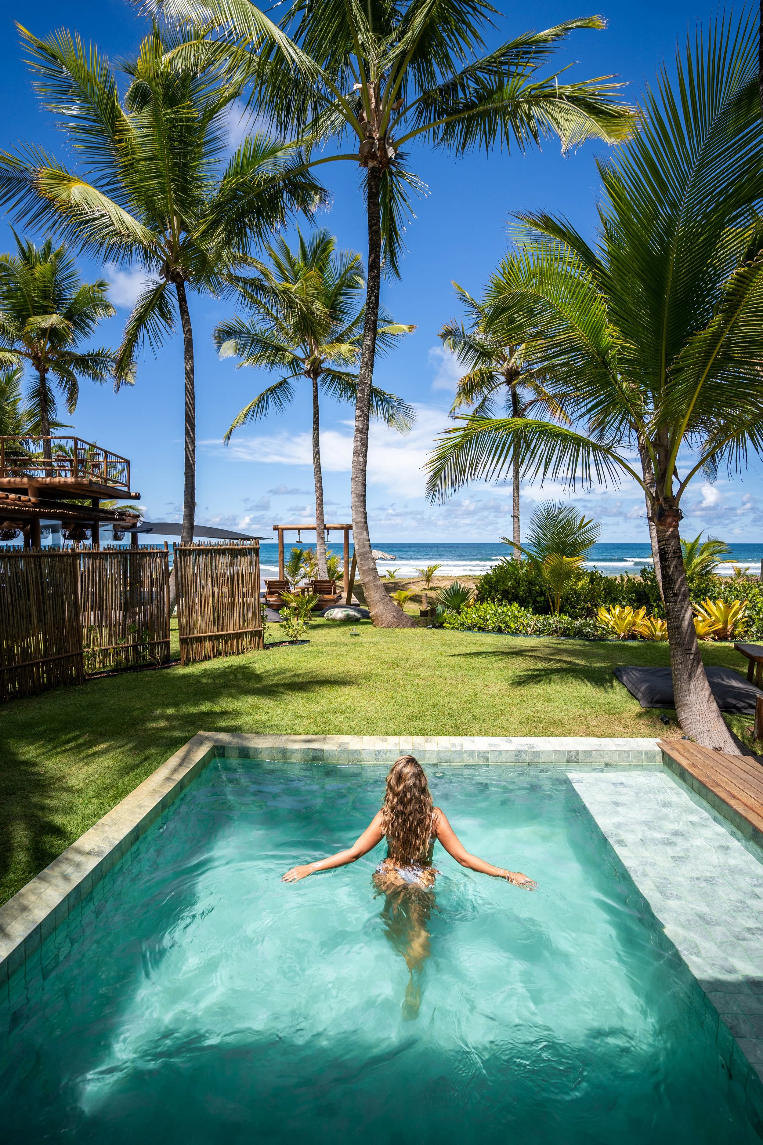 Mulher em piscina com vista para palmeiras e praia ao fundo em um cenário tropical ensolarado.