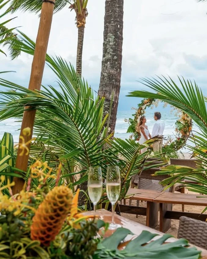 Cena de casamento na praia com decoração tropical, incluindo palmeiras e flores, duas taças de champanhe em primeiro plano e um casal em pé próximo ao arco decorativo, com o mar ao fundo.
