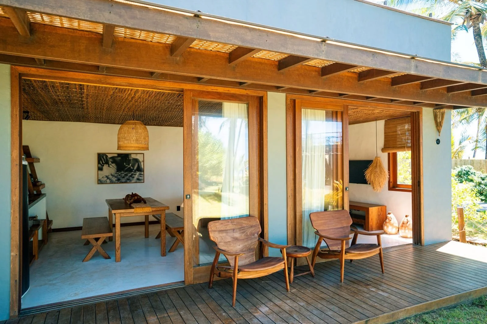 Varanda com duas cadeiras de madeira e uma mesa ao ar livre. Interior de uma casa de madeira com móveis em tom natural, decoração de estilo rústico e janelas grandes com cortinas leves, mostrando um ambiente de descanso.