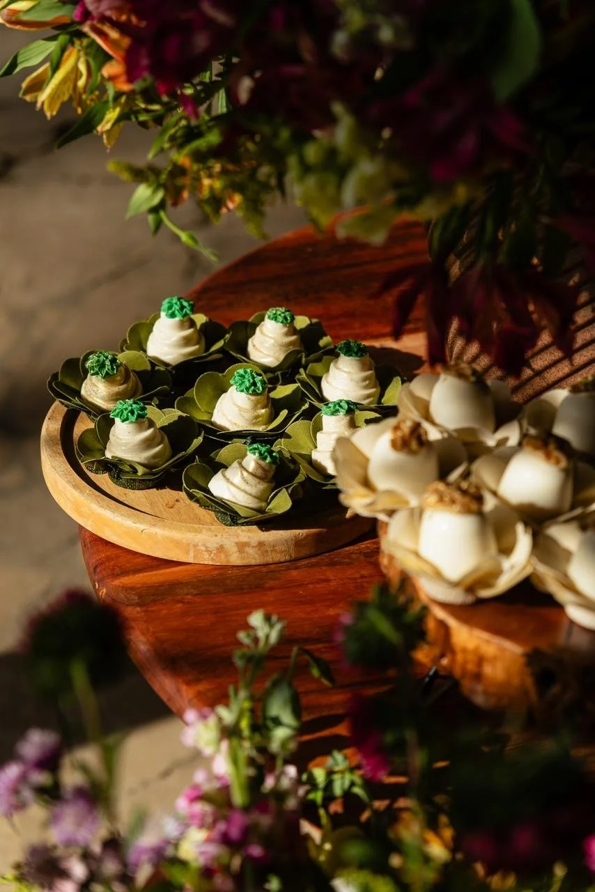 Doces decorativos brancos e verdes dispostos em folhas verdes em uma mesa de madeira, em um ambiente decorado com flores.